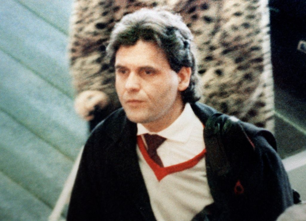 A man with dark hair wearing a suit and tie walks indoors, partially obscured by someone in a patterned coat