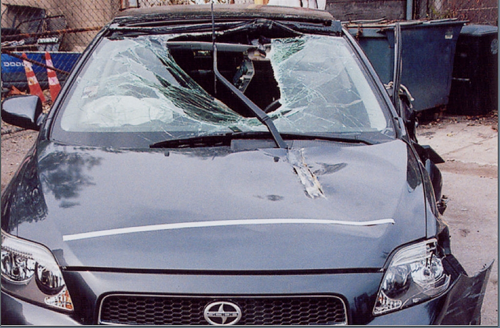 Damaged car with shattered windshield and bent roof, parked near dumpsters in an alleyway