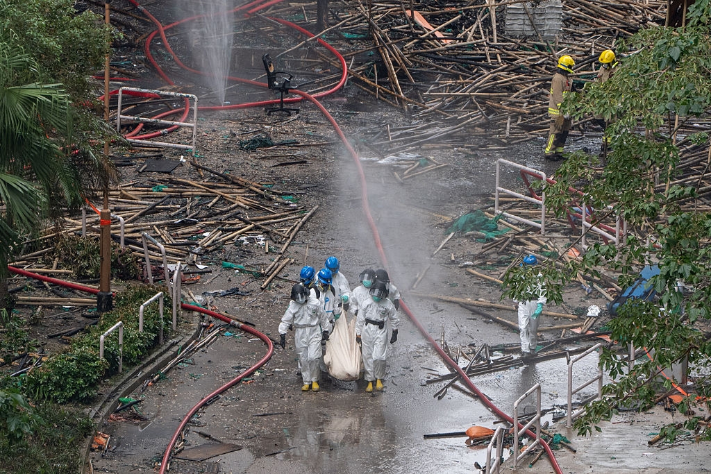 Emergency workers in protective suits carry a bag through debris at a disaster site, with hoses and scattered materials in the background