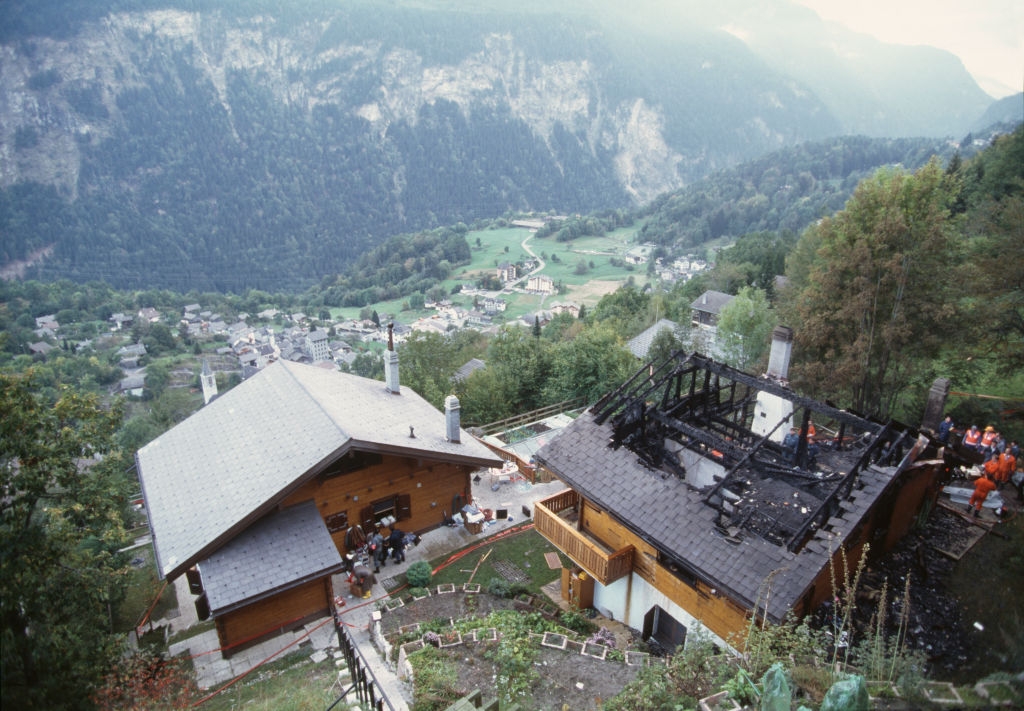 Aerial view of a burned-out house next to a standing house, set in a mountainous village with trees and valley in the background