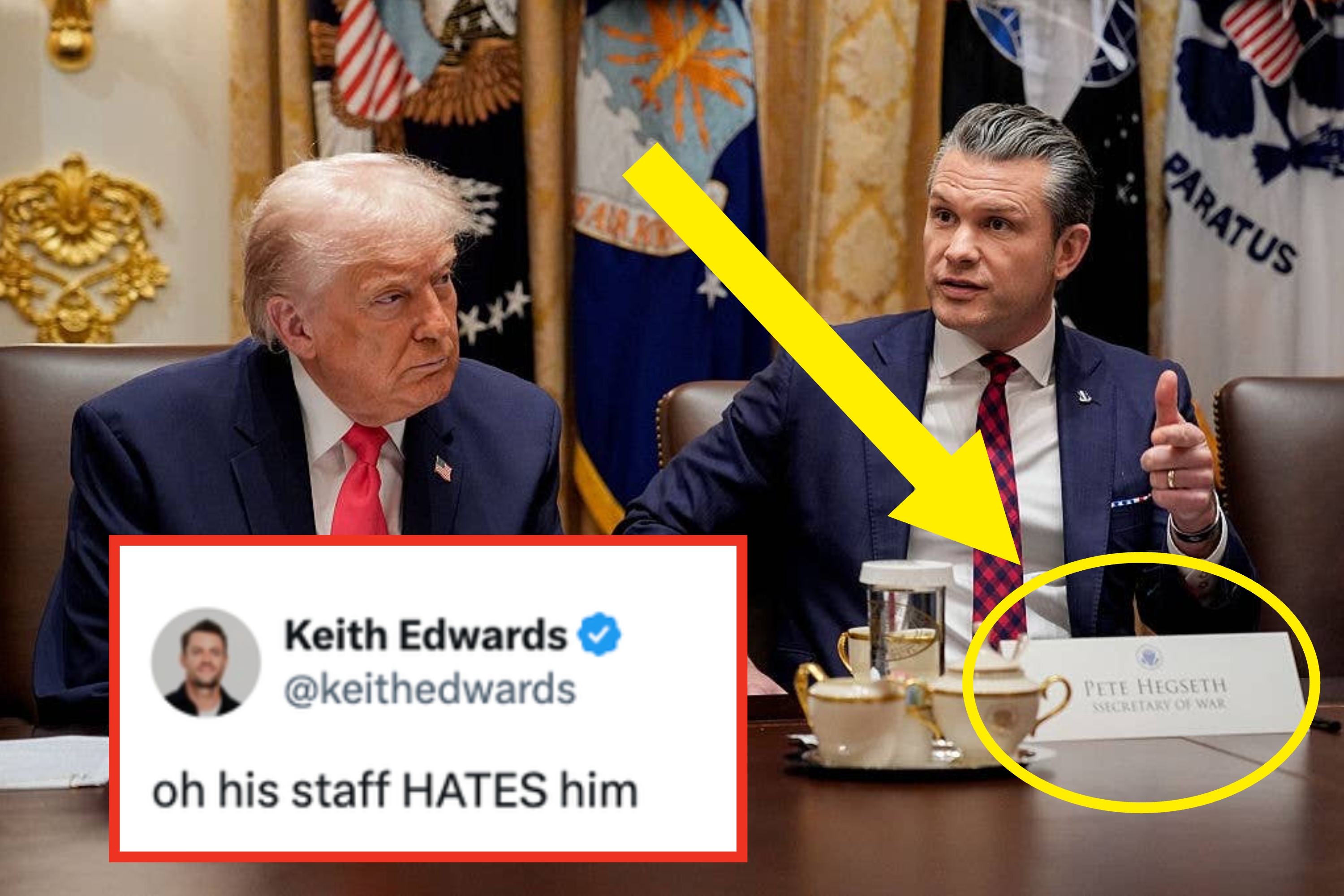 A man in a suit with a red patterned tie sits at a conference table, speaking, with another man in a suit listening beside him