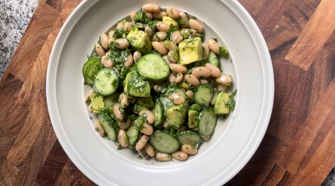 A bowl of salad with sliced cucumber, avocado, white beans, and chopped herbs on a wooden table
