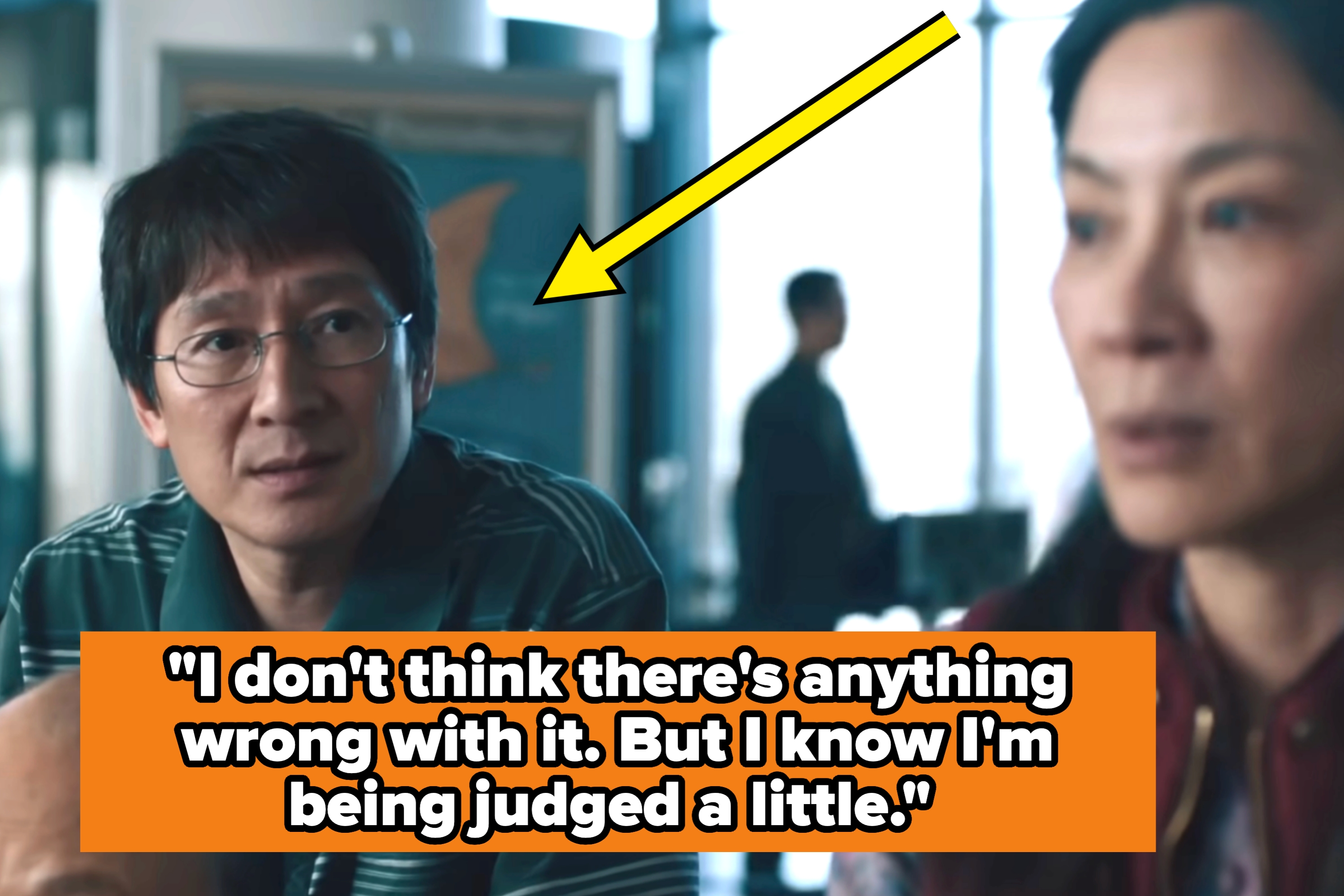 A man and woman are talking indoors with a blurry figure in the background. The man looks towards the woman, appearing to wear casual attire