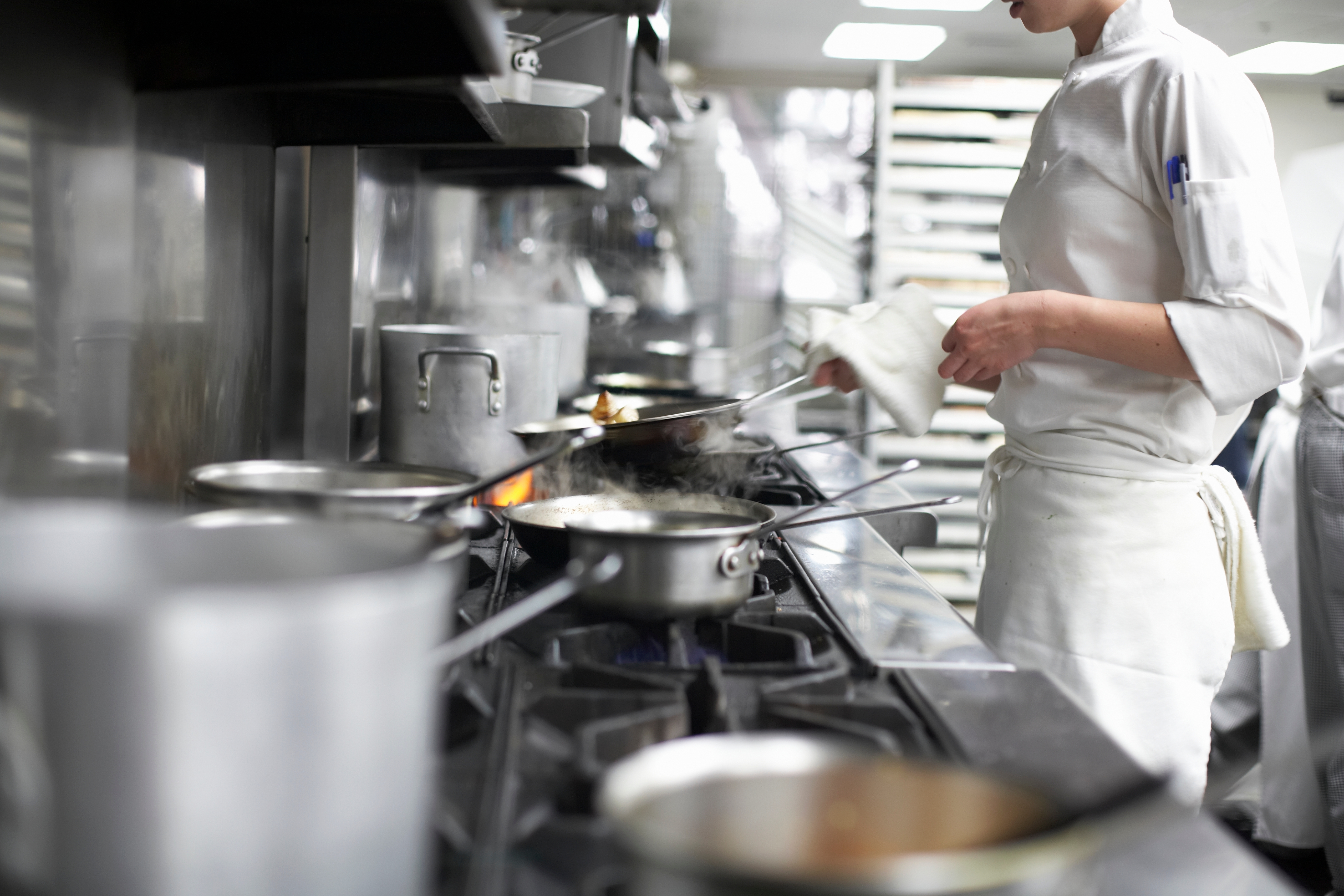 Chef in a busy restaurant kitchen, wearing a white uniform, cooking multiple dishes over a stove with steaming pots