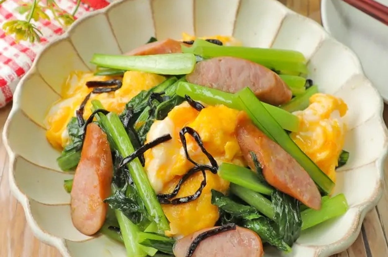 スクランブルエッグ、ソーセージ、小松菜、ひじきを使った料理が皿に盛られている。木製のテーブルに置かれている。