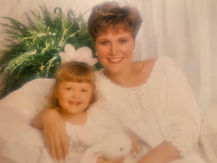 Woman and child in white outfits smile while seated on a couch with a plush toy and a leafy plant in the background