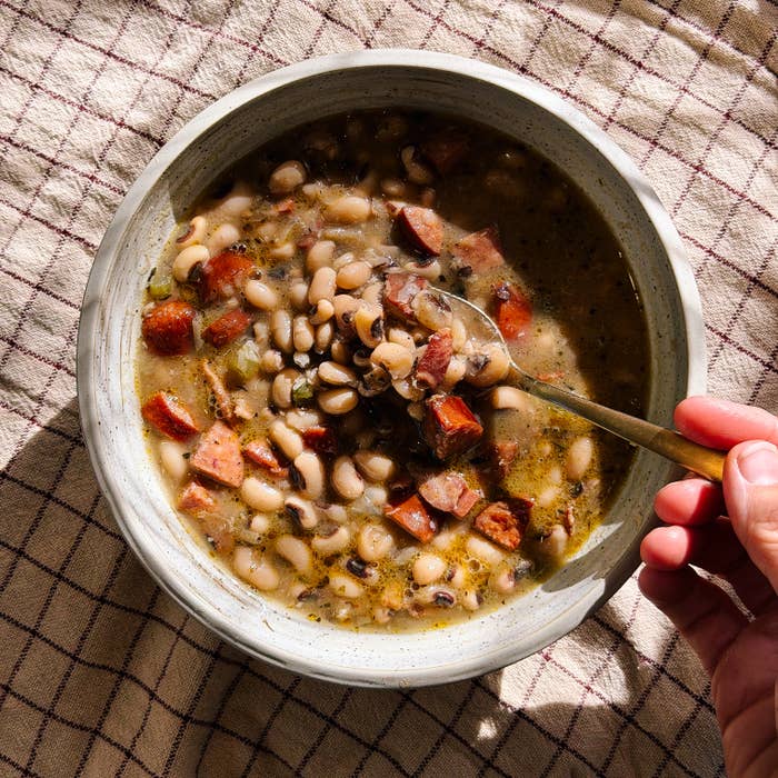 A bowl of bean soup with chunks of meat, being stirred by a hand holding a spoon on a cloth background