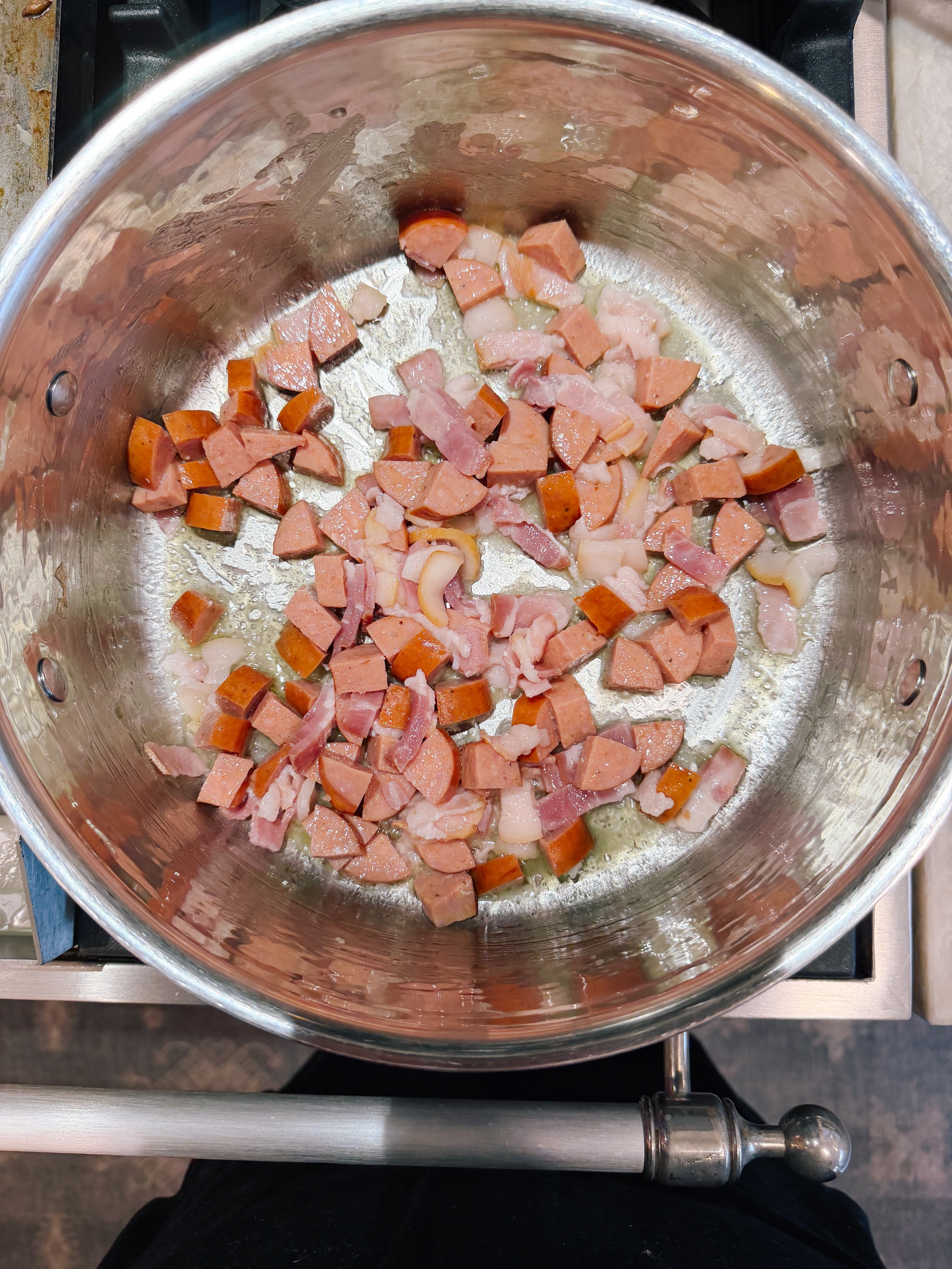 Diced sausages and bacon cook in a large pot, beginning to brown