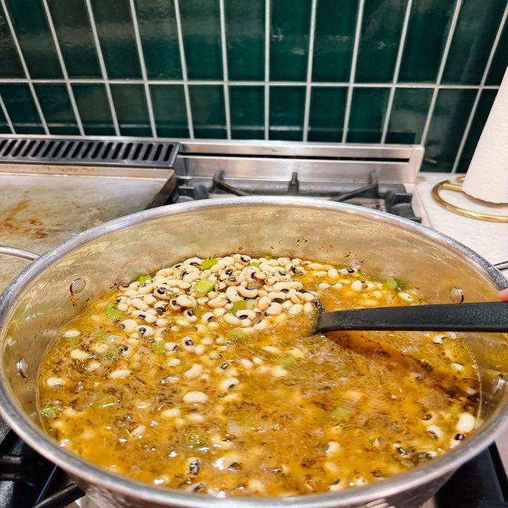 A pot on a stove with soup being stirred, containing beans and herbs, set in a tiled kitchen