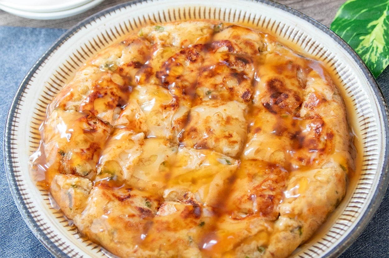 皿に盛られたれんこんチヂミ。あんがかかっていて、おいしそうな見た目。料理の背景には緑の葉がある。