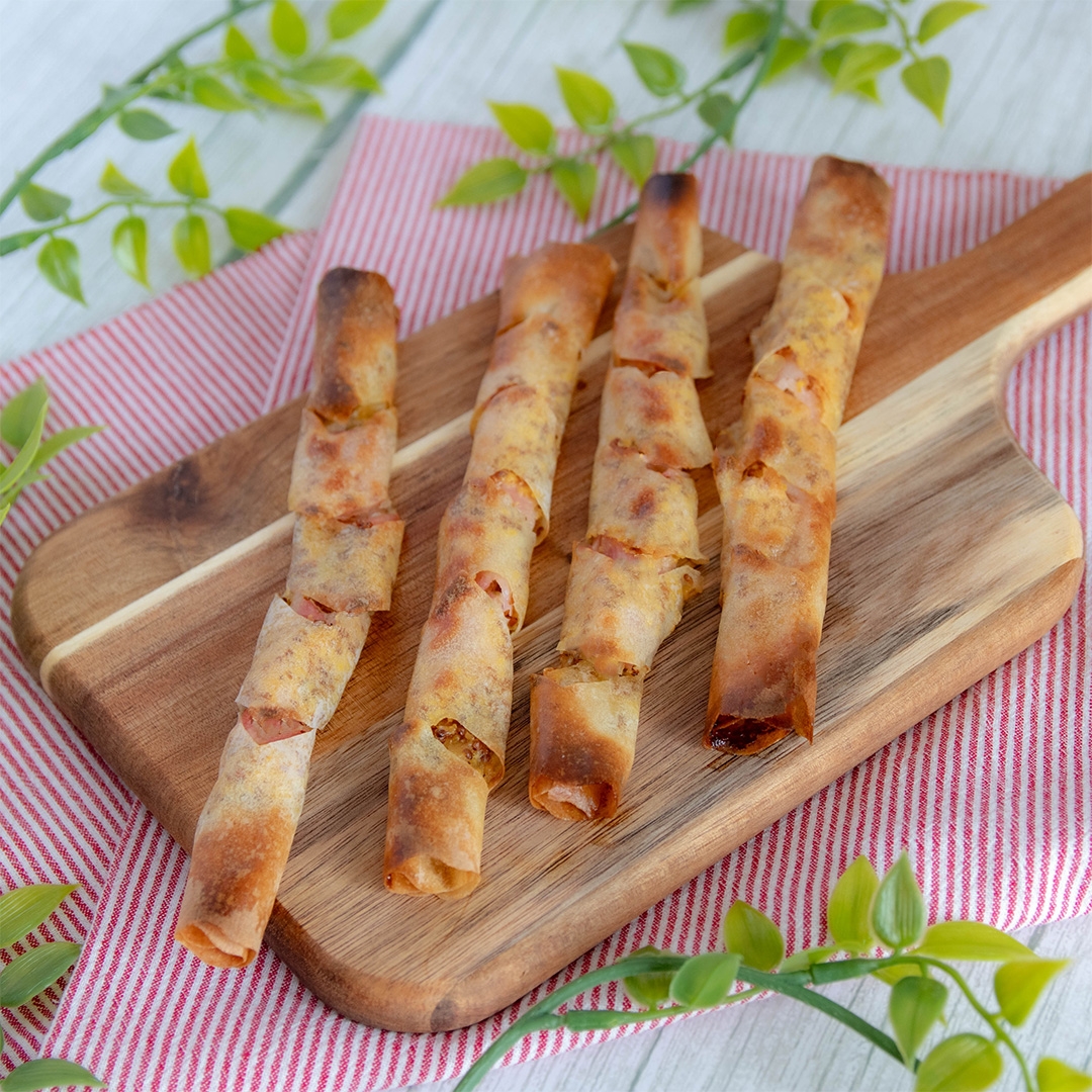 カットボードに細長い春巻き料理が4本置かれ、周囲に緑の葉が点在しています。