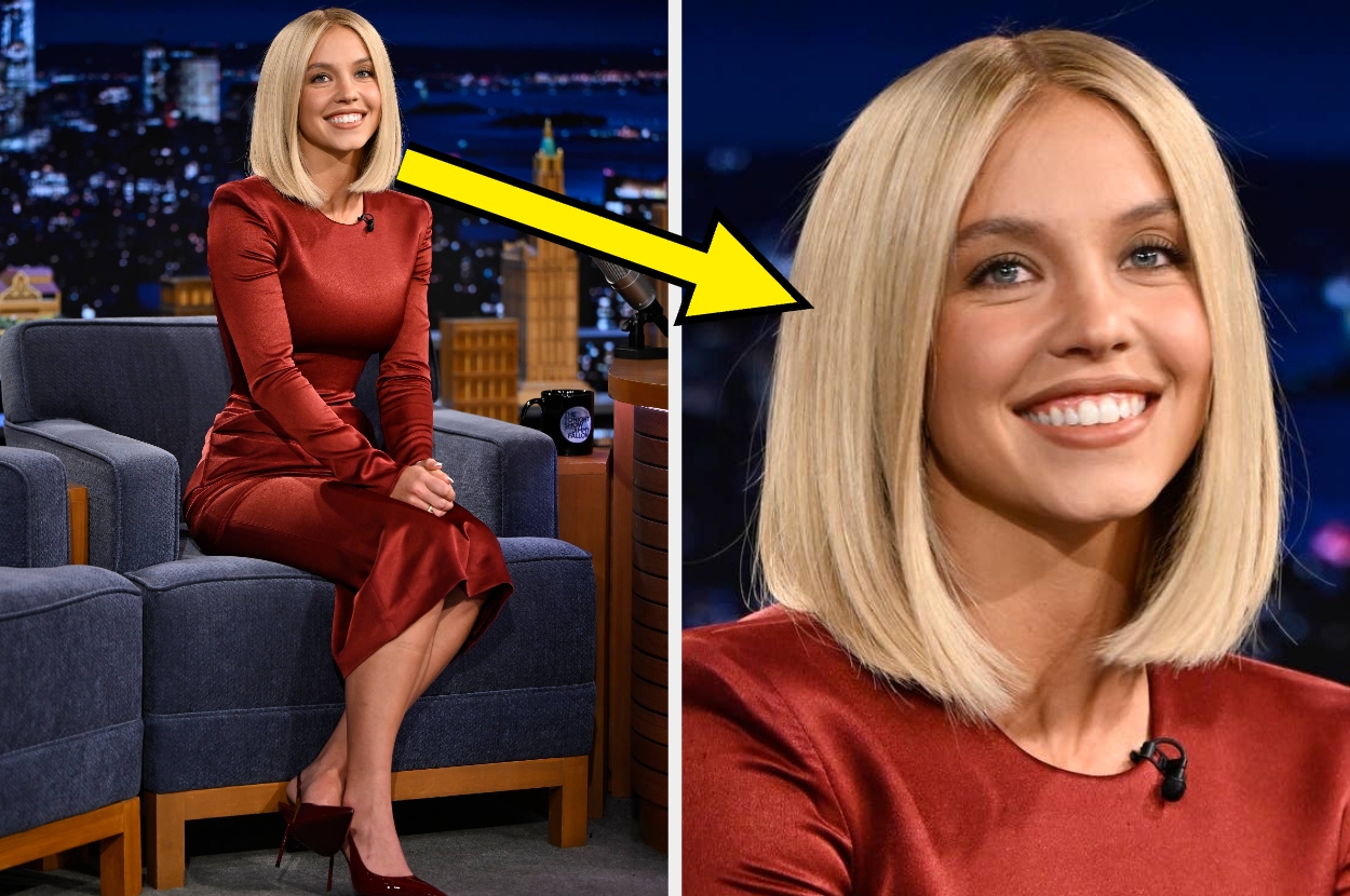 Person on a talk show set, smiling and wearing a sleek, fitted red dress, seated in front of a cityscape backdrop