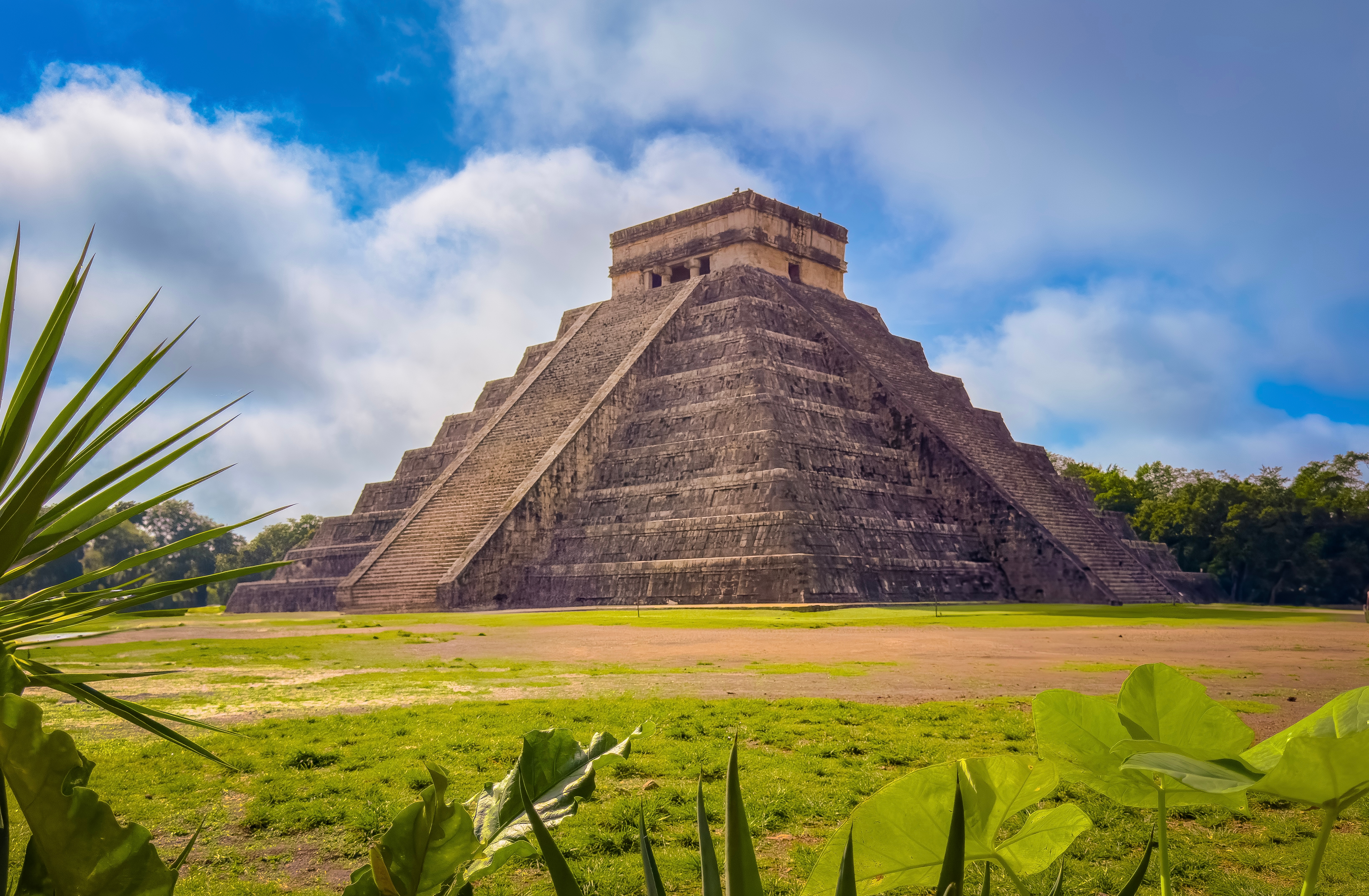 Chichén Itzá pyramid stands against a cloudy sky, surrounded by greenish foliage and a grassy foreground