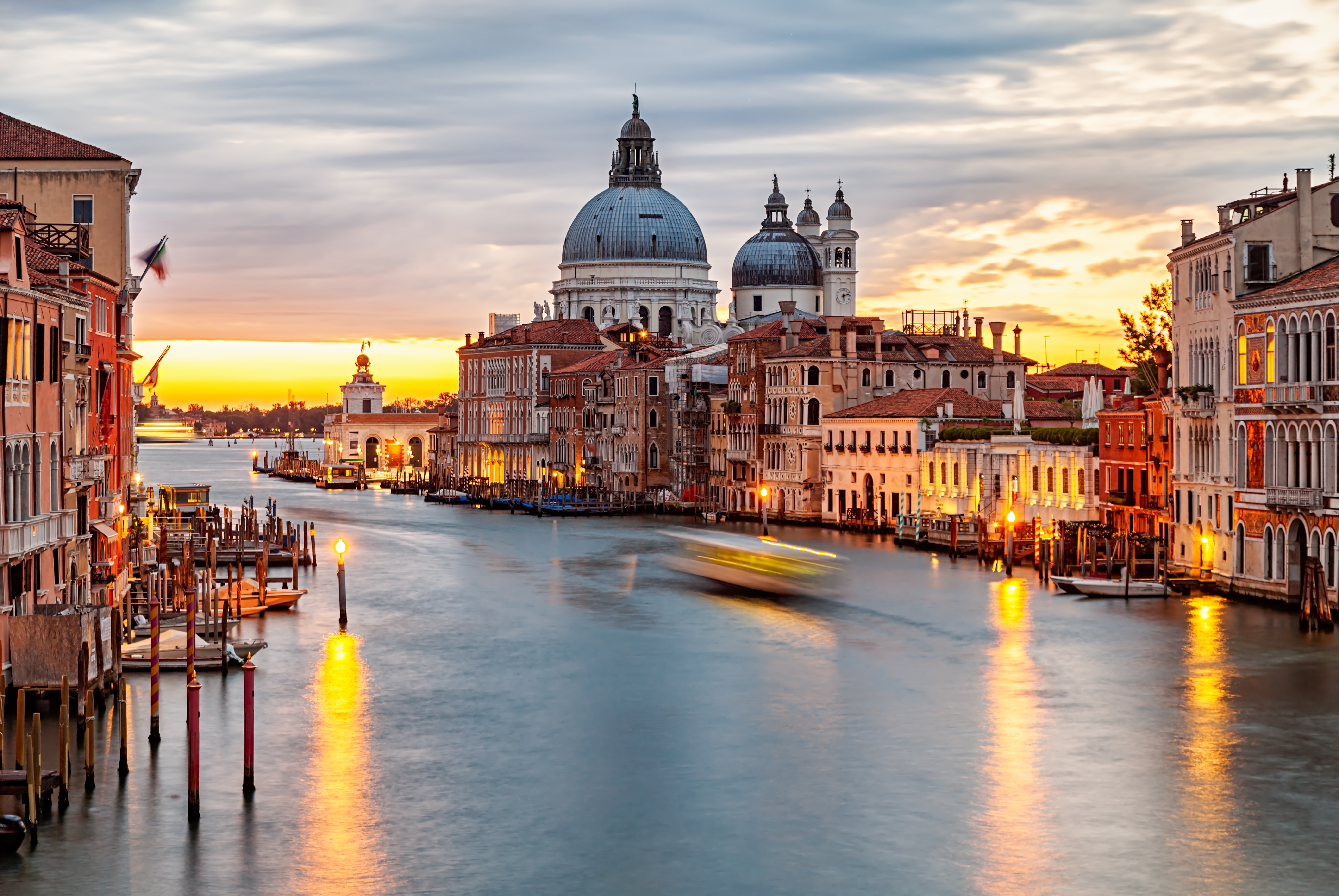 Scenic presumption of Venice's Grand Canal astatine sunset, featuring historical architecture and a distant basilica dome nether a partially cloudy sky