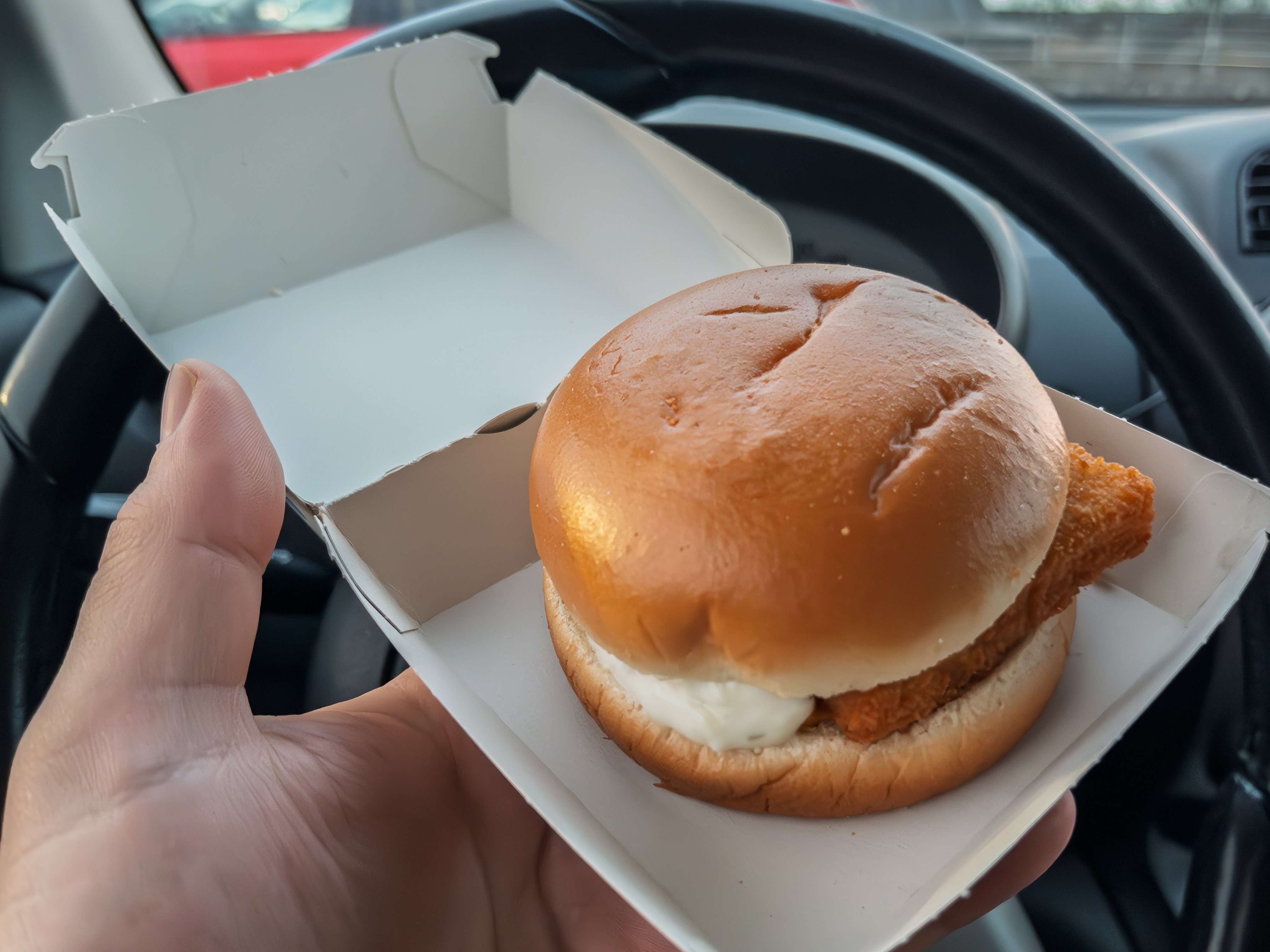 Fish sandwich in a box held up inside a car, with steering wheel in the background