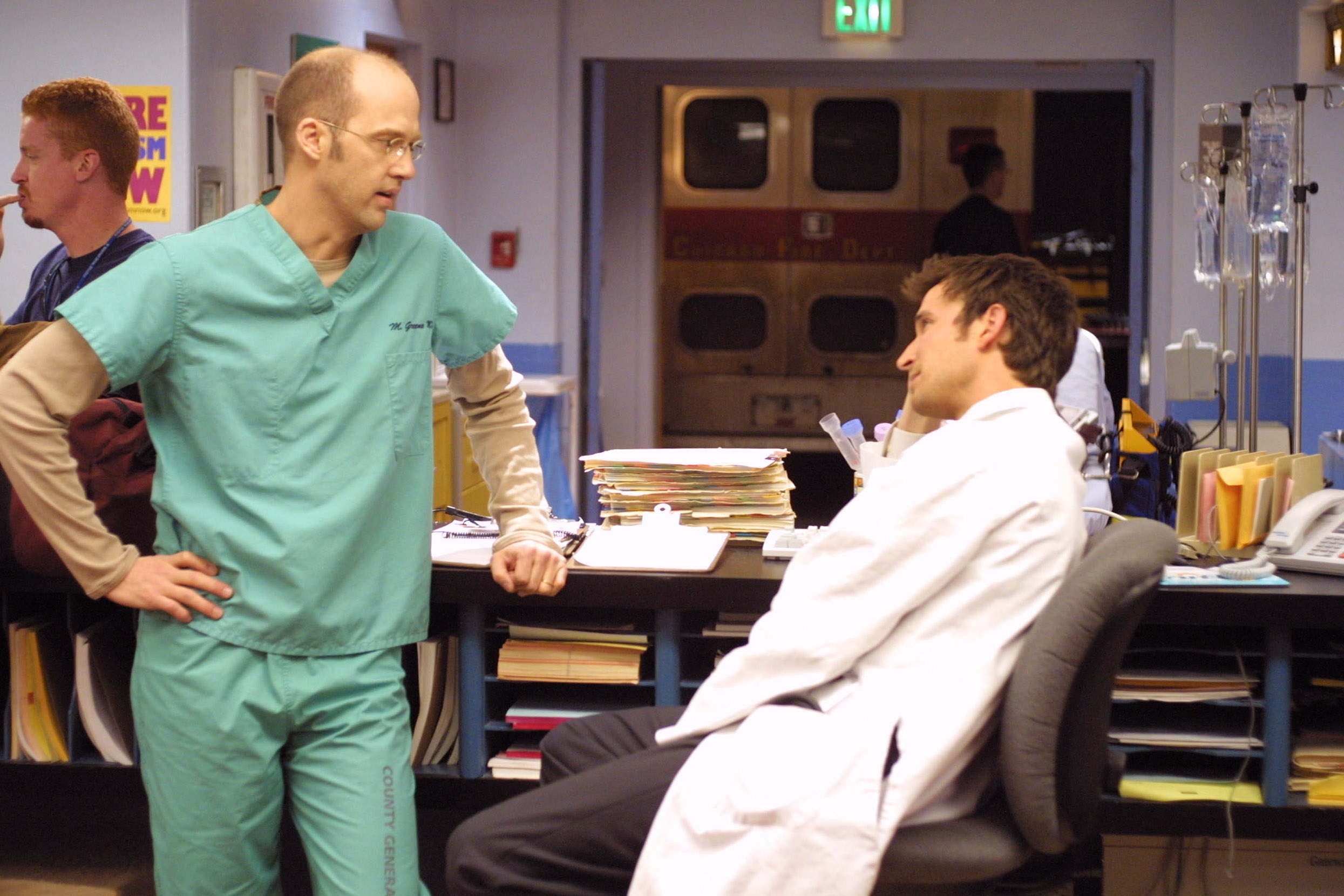  one in scrubs, the other in a lab coat, talking near a busy nurse's station