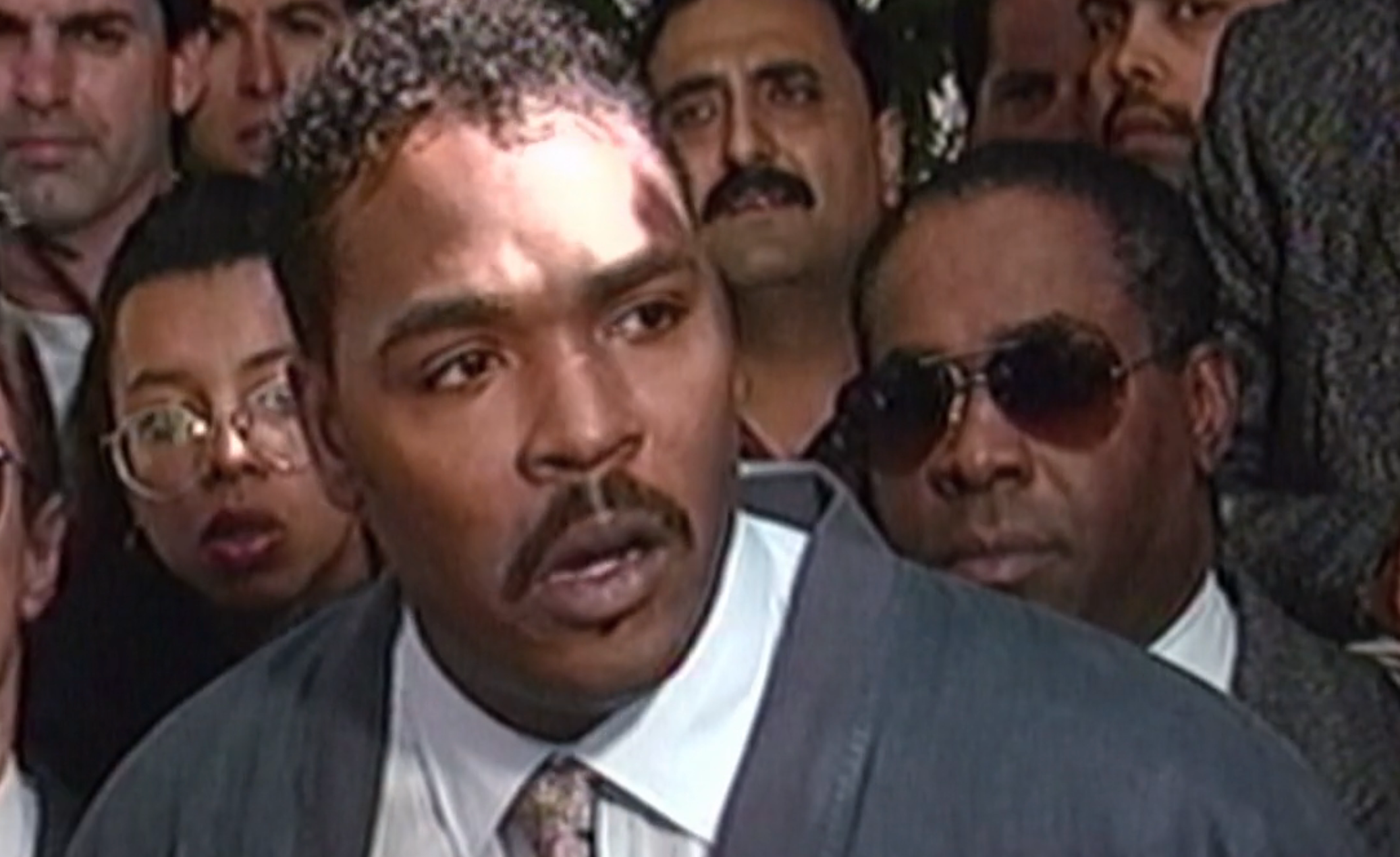 I don't know who this person is, but they are speaking to the press surrounded by people, wearing a suit jacket, shirt, and tie