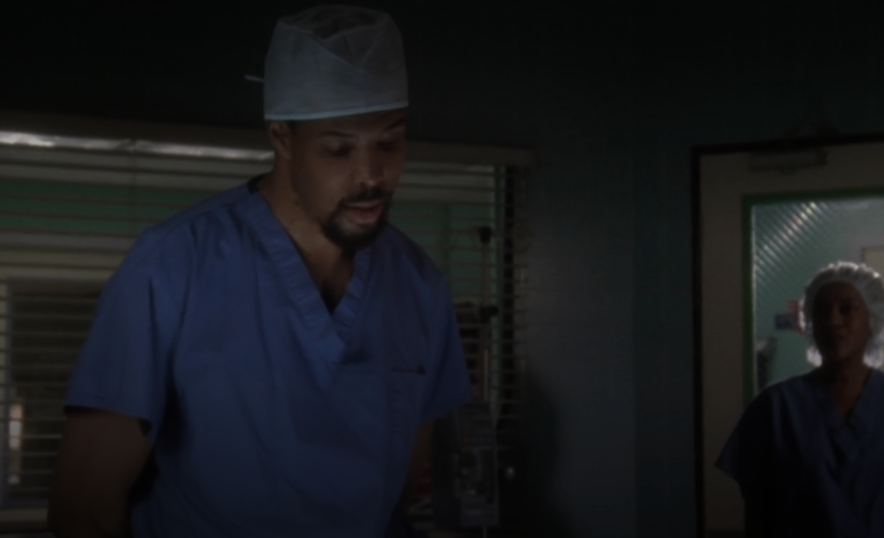 Two people in surgical attire inside a dimly lit hospital room, appearing to discuss or perform a medical procedure