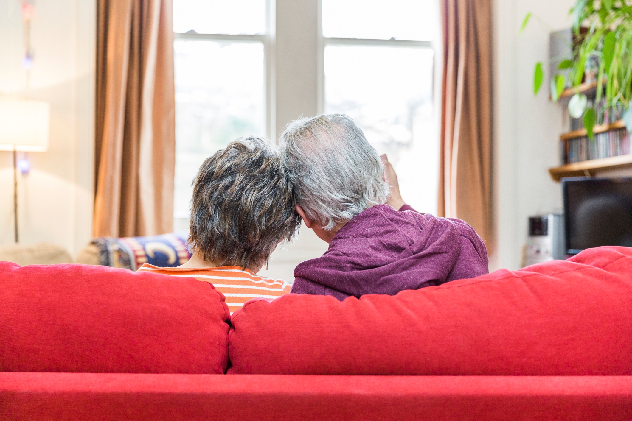 Et eldre par sitter tett på en sofa, lener hodene sammen, sett bakfra, i et koselig rom med et vindu i bakgrunnen