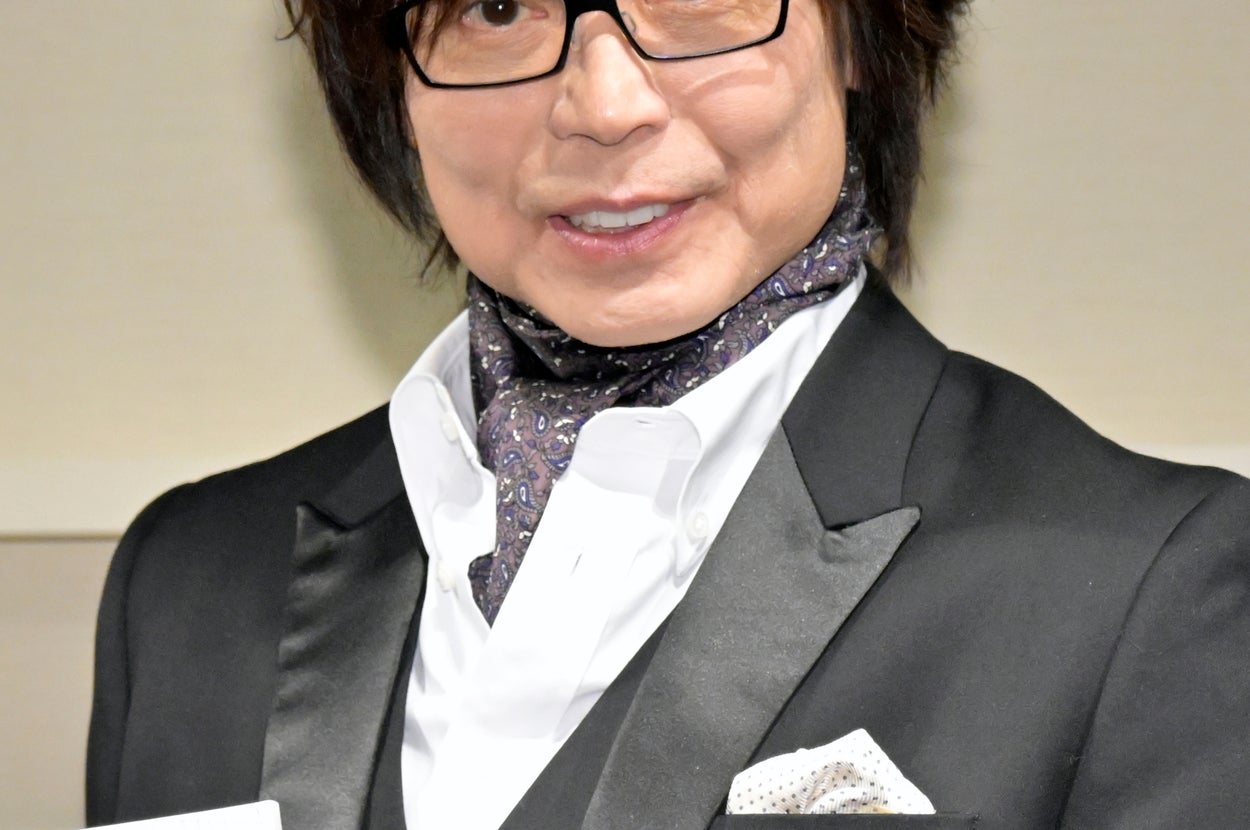 有名人がフォーマルな黒いスーツを着て、スカーフを巻き、四角いオブジェクトを持っています。