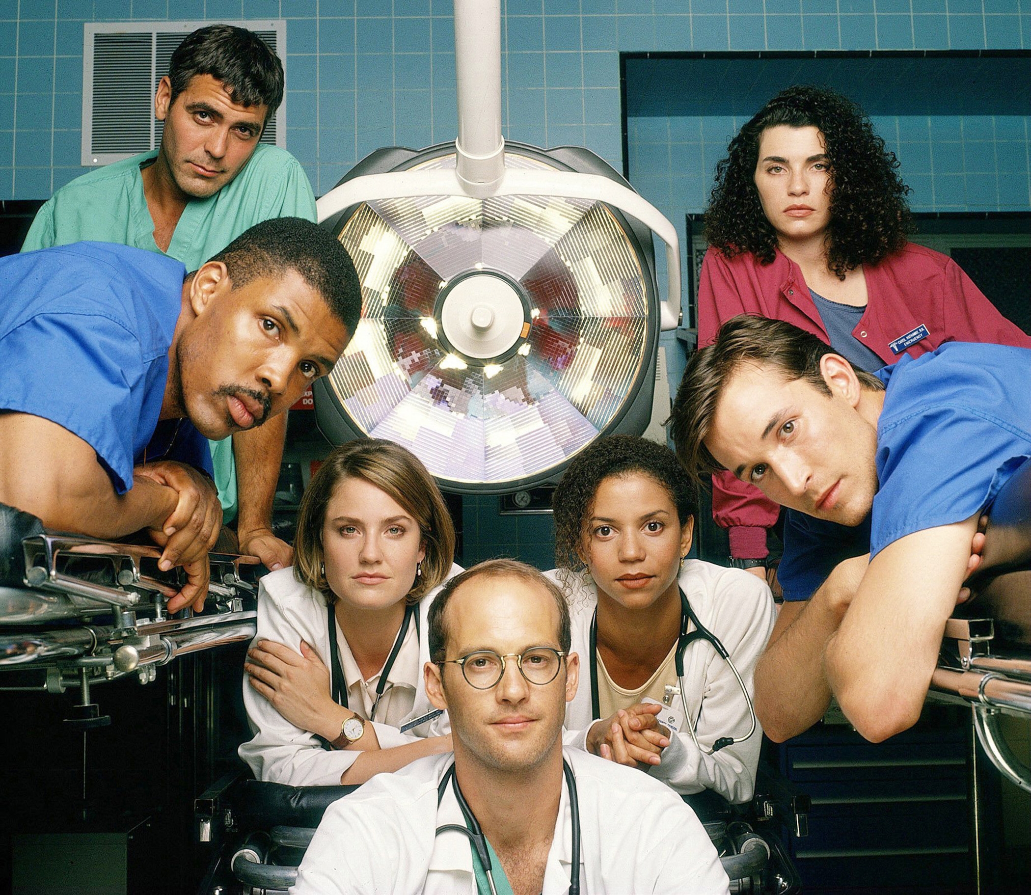 Group of actors in a medical drama setting, wearing scrubs and stethoscopes, gathered around a surgery table