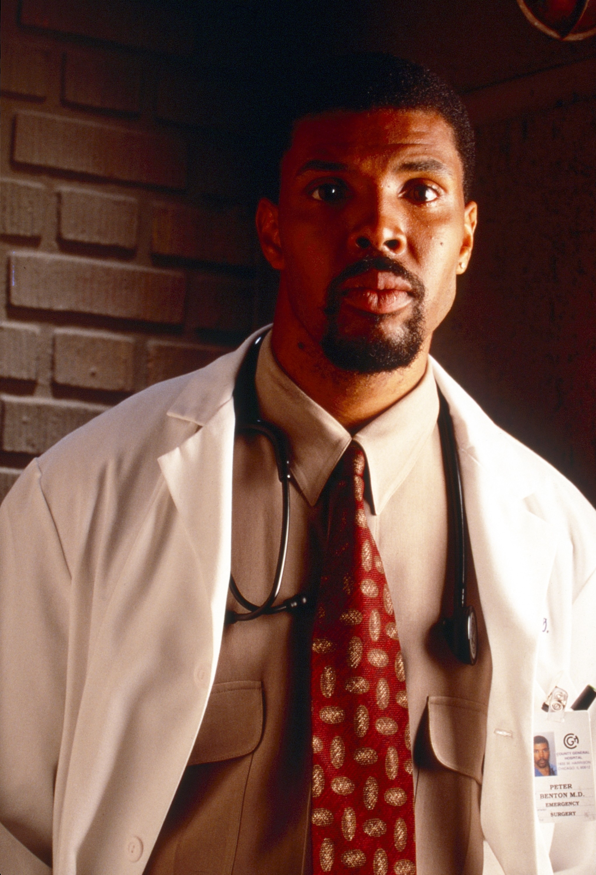 Actor in a medical drama scene wearing a beige shirt, red patterned tie, and a white lab coat with a stethoscope around the neck