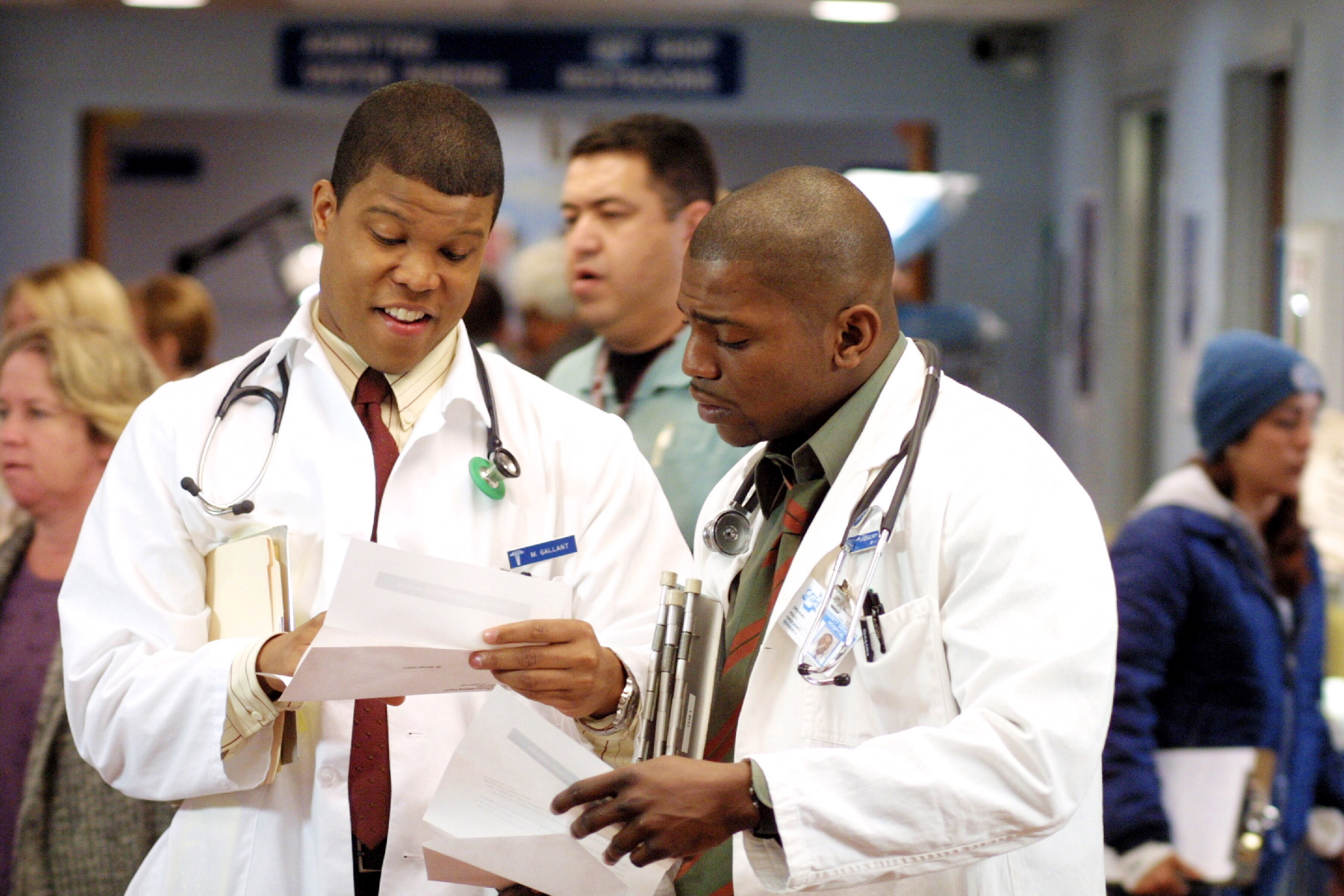 Two doctors in lab coats review papers in a bustling hospital setting. People are visible in the background, suggesting a busy environment