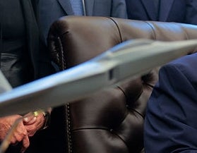 En gruppe mennesker står rundt en person som sitter ved et skrivebord med en jetmodell og Abraham Lincoln-byste i forgrunnen