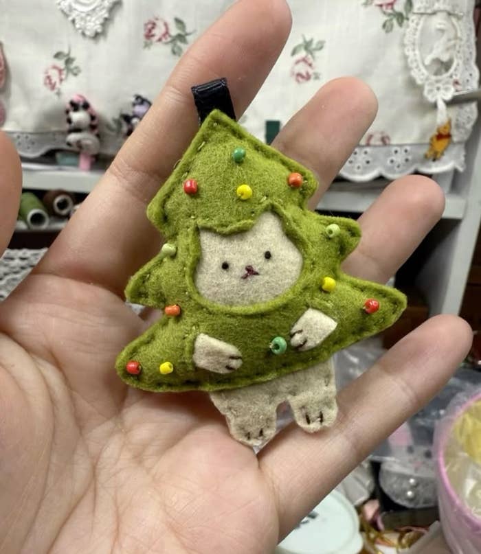 Adorno navideño de fieltro en forma de árbol con un diseño de gato en el centro, sostenido en la palma de una mano