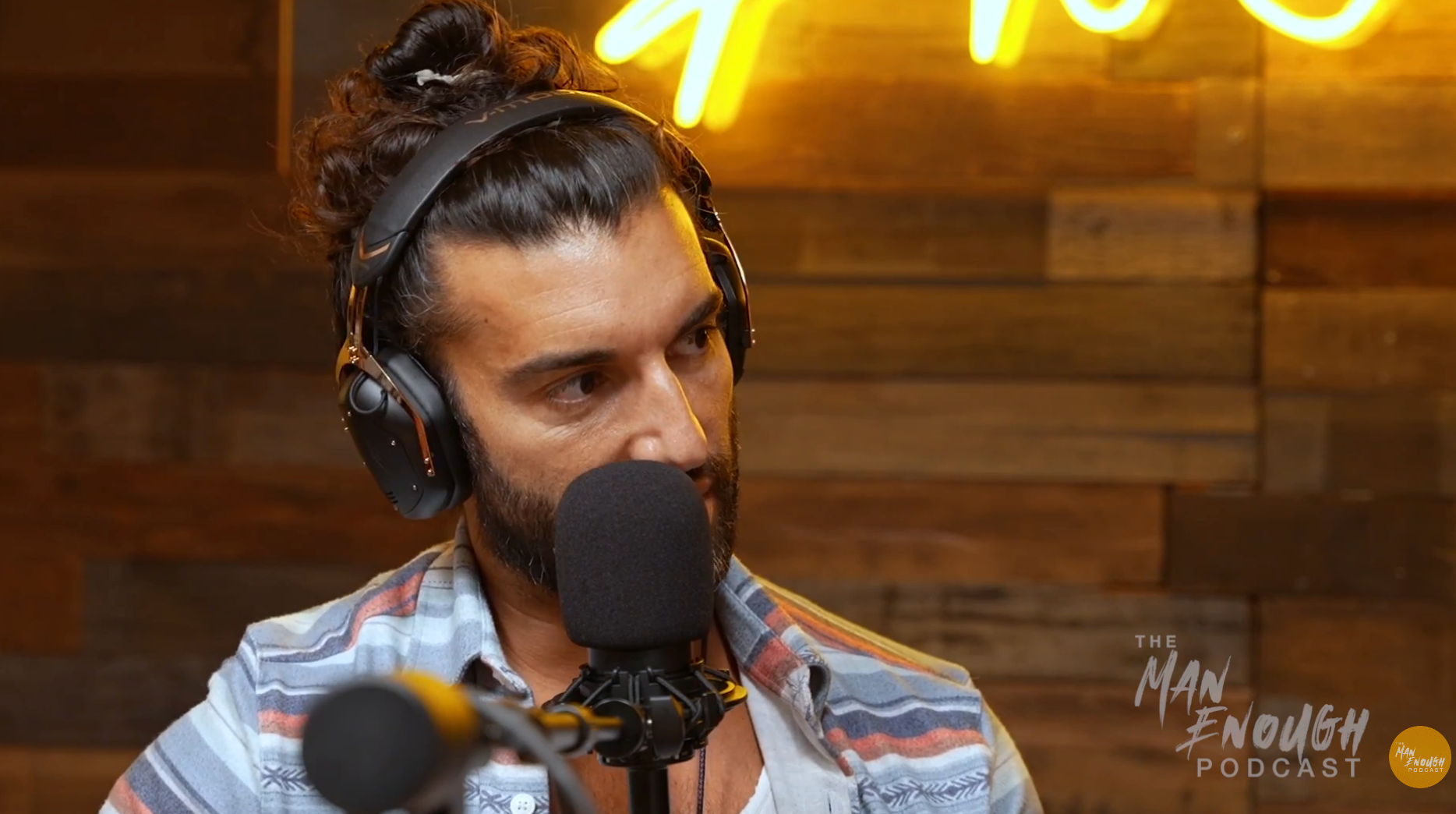 Person with headphones speaking into a microphone during a podcast recording, with logo "The Man Enough Podcast" visible in the background