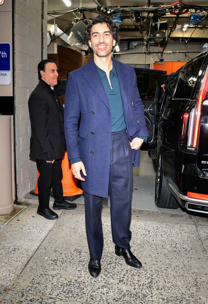 Person in a dark coat and dress shoes stands near a vehicle, smiling, with a security guard in the background