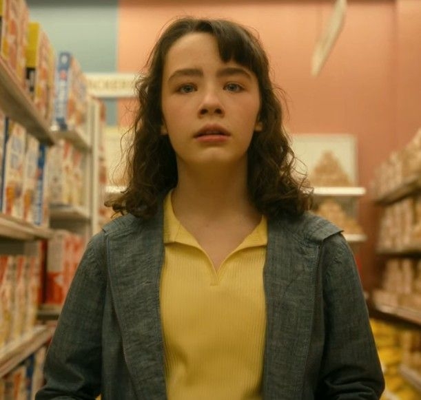 Persona joven con chaqueta de mezclilla y camisa amarilla, mirando al frente en el pasillo de un supermercado. Fondo de estantes con cajas