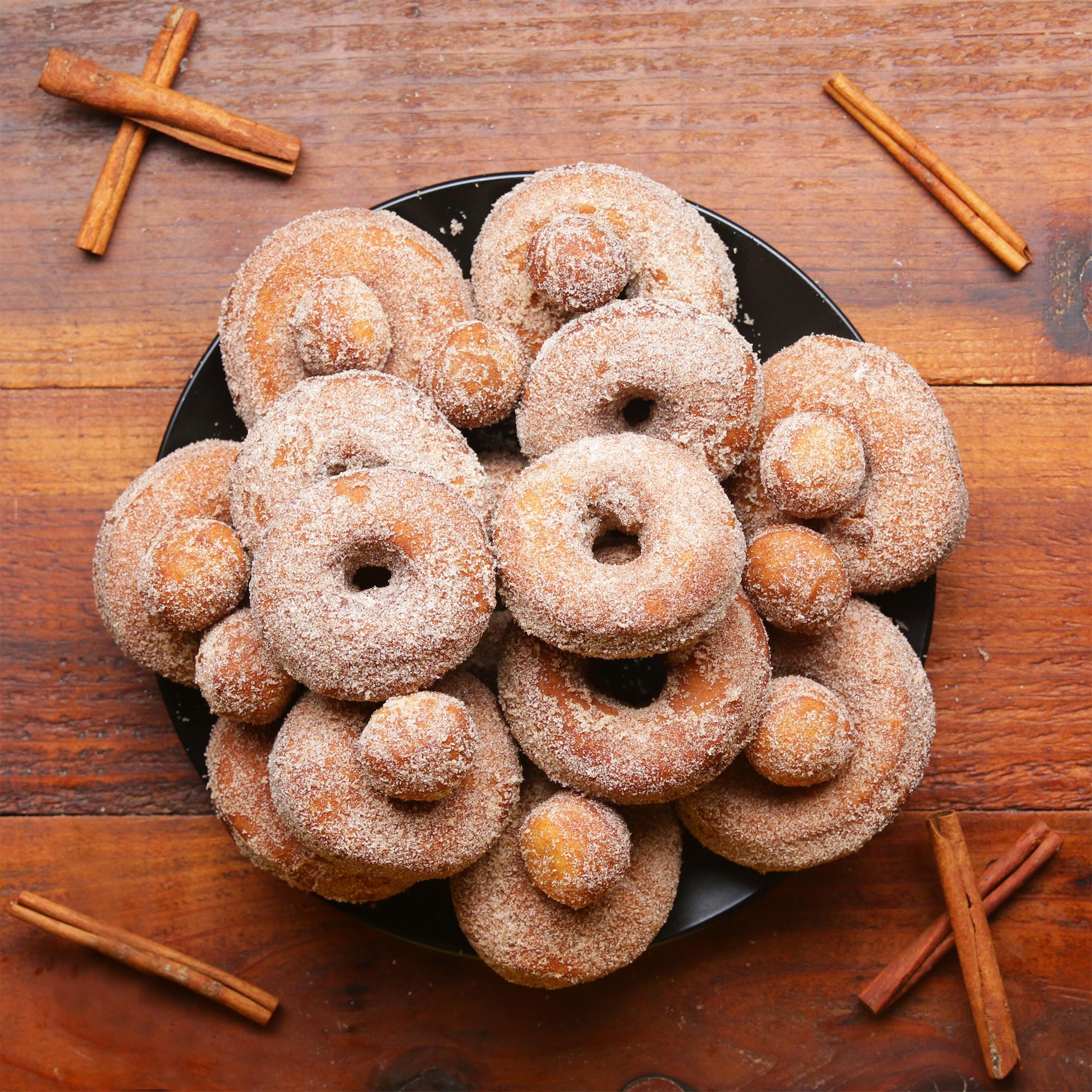A plate piled with sugar-coated donuts, surrounded by cinnamon sticks on a wooden table