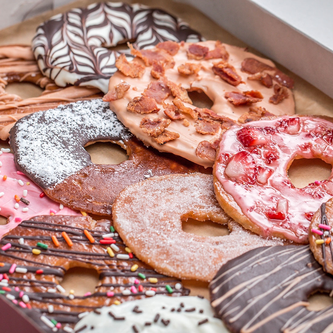 Assorted gourmet donuts with toppings like bacon, sprinkles, powdered sugar, chocolate drizzle, and fruit glaze arranged in a box