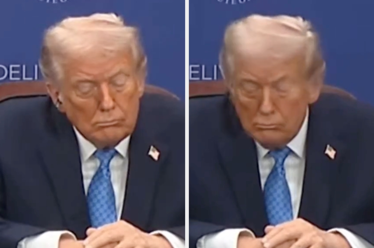 A person at a conference table with hands clasped, wearing a suit and blue tie, eyes closed, in a focused or contemplative posture