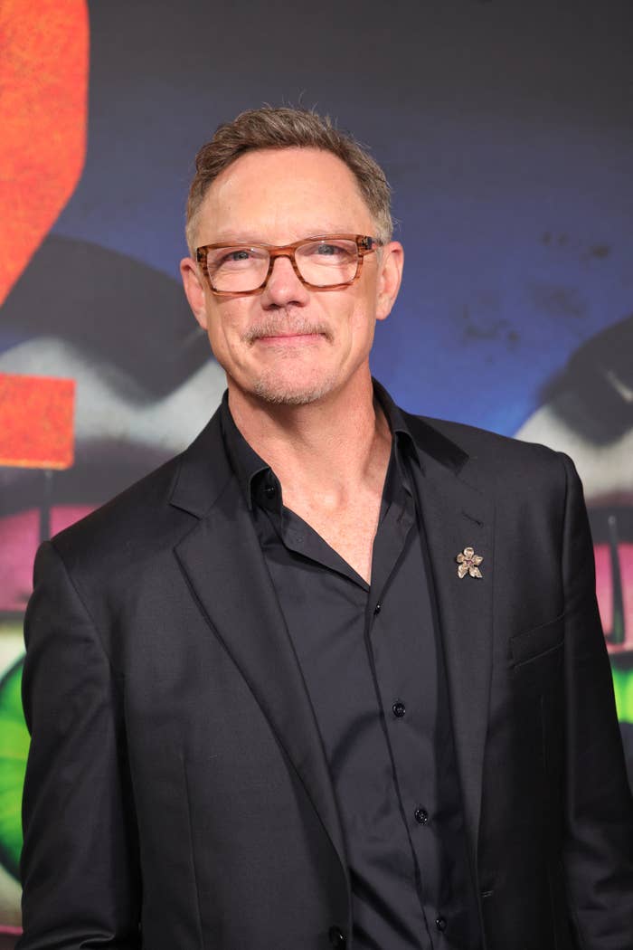 Matthew Lillard in a black suit and shirt with glasses at a public event, standing in front of a vibrant, graffiti-style backdrop