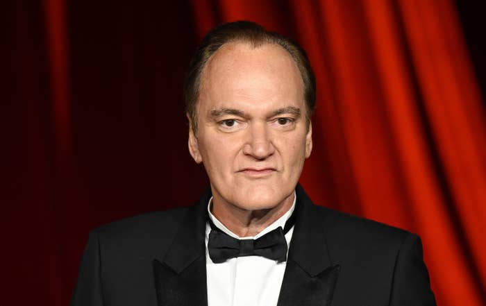 Quentin Tarantino in a formal black tuxedo poses in front of a red curtain backdrop