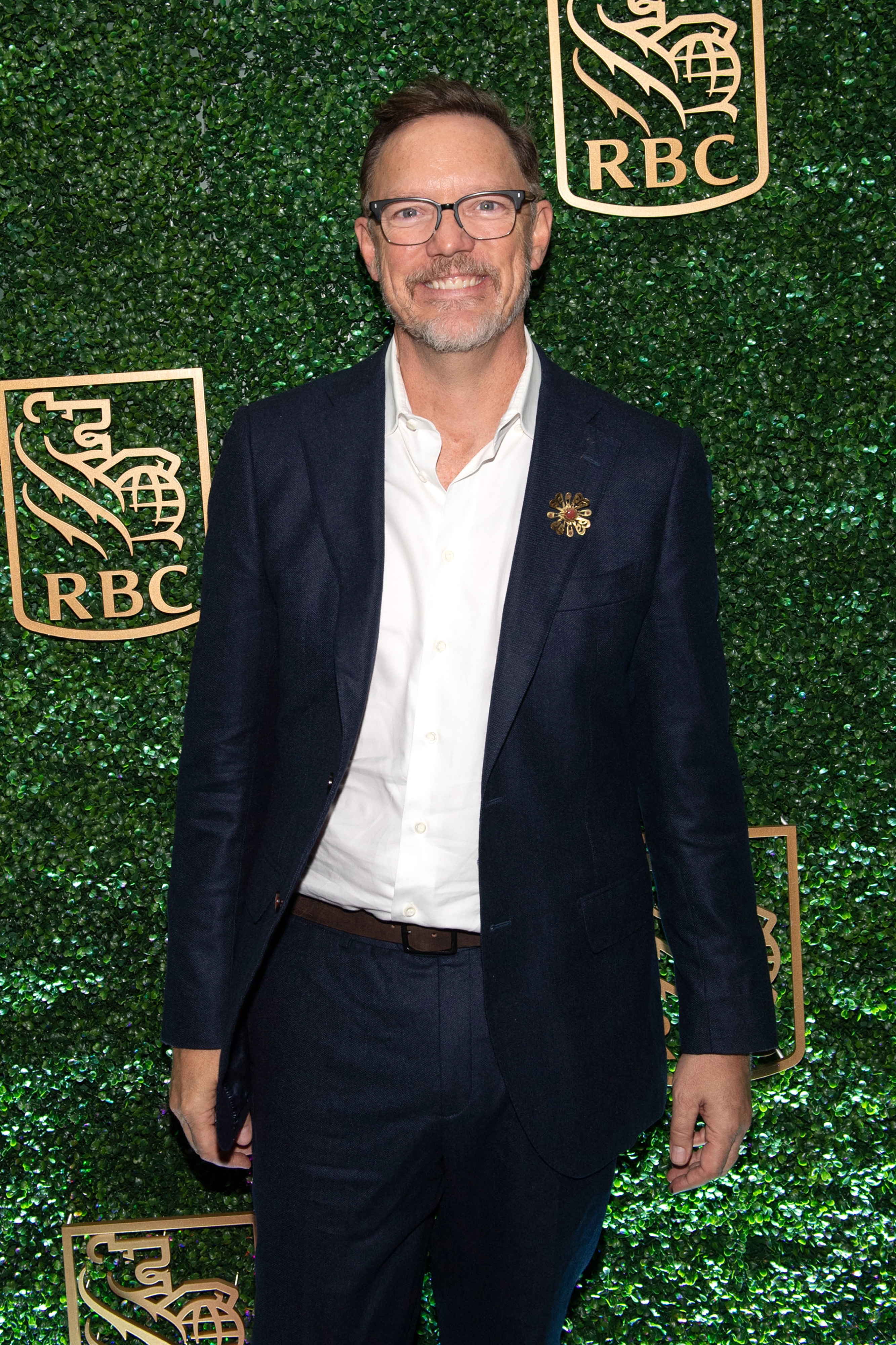 Matthew Lillard in a suit and open-collared shirt poses in front of a greenery backdrop with an RBC logo