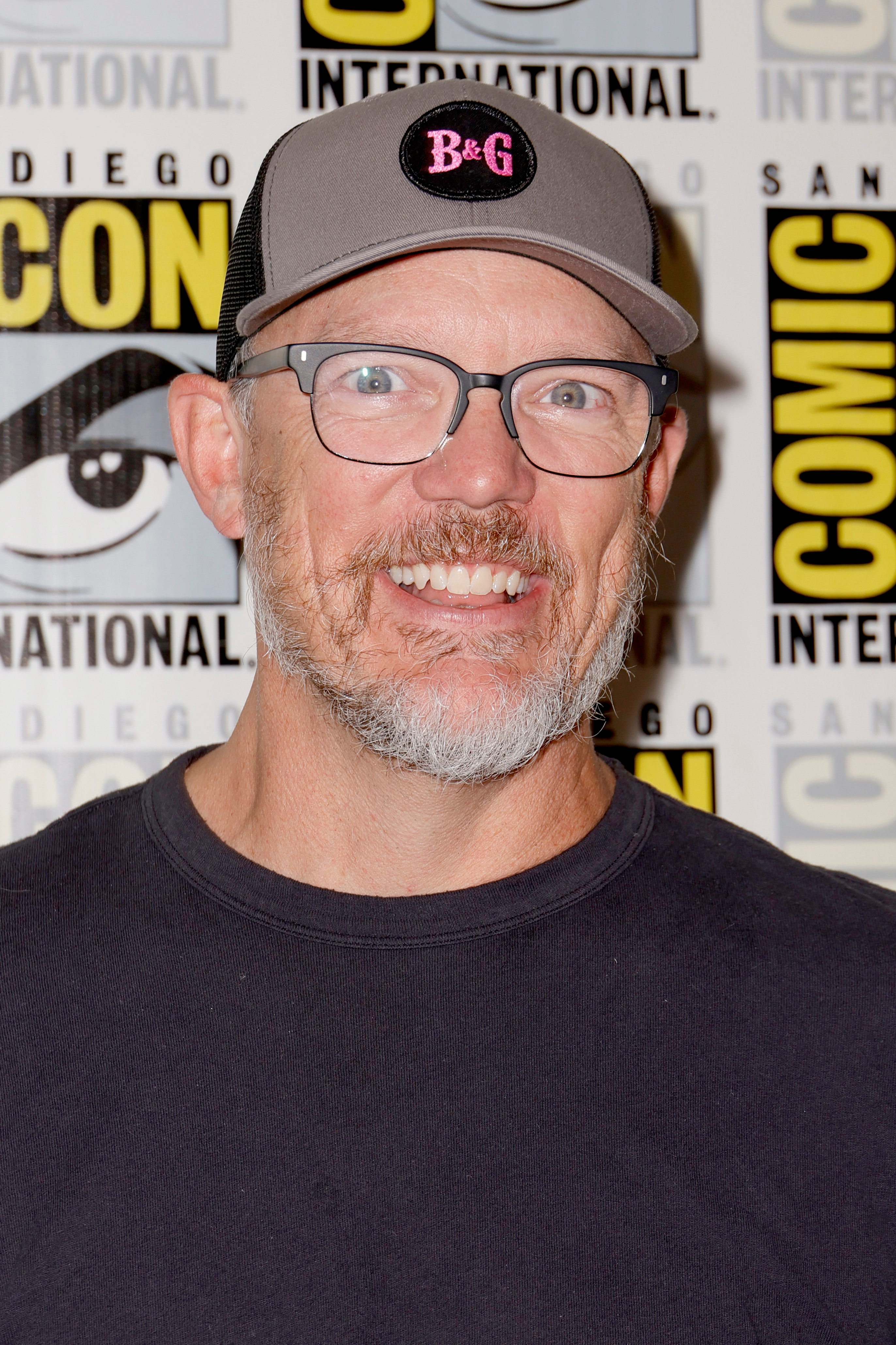 Matthew Lillard smiling at a Comic-Con event, wearing glasses, a black shirt, and a cap. Comic-Con logos are visible in the background