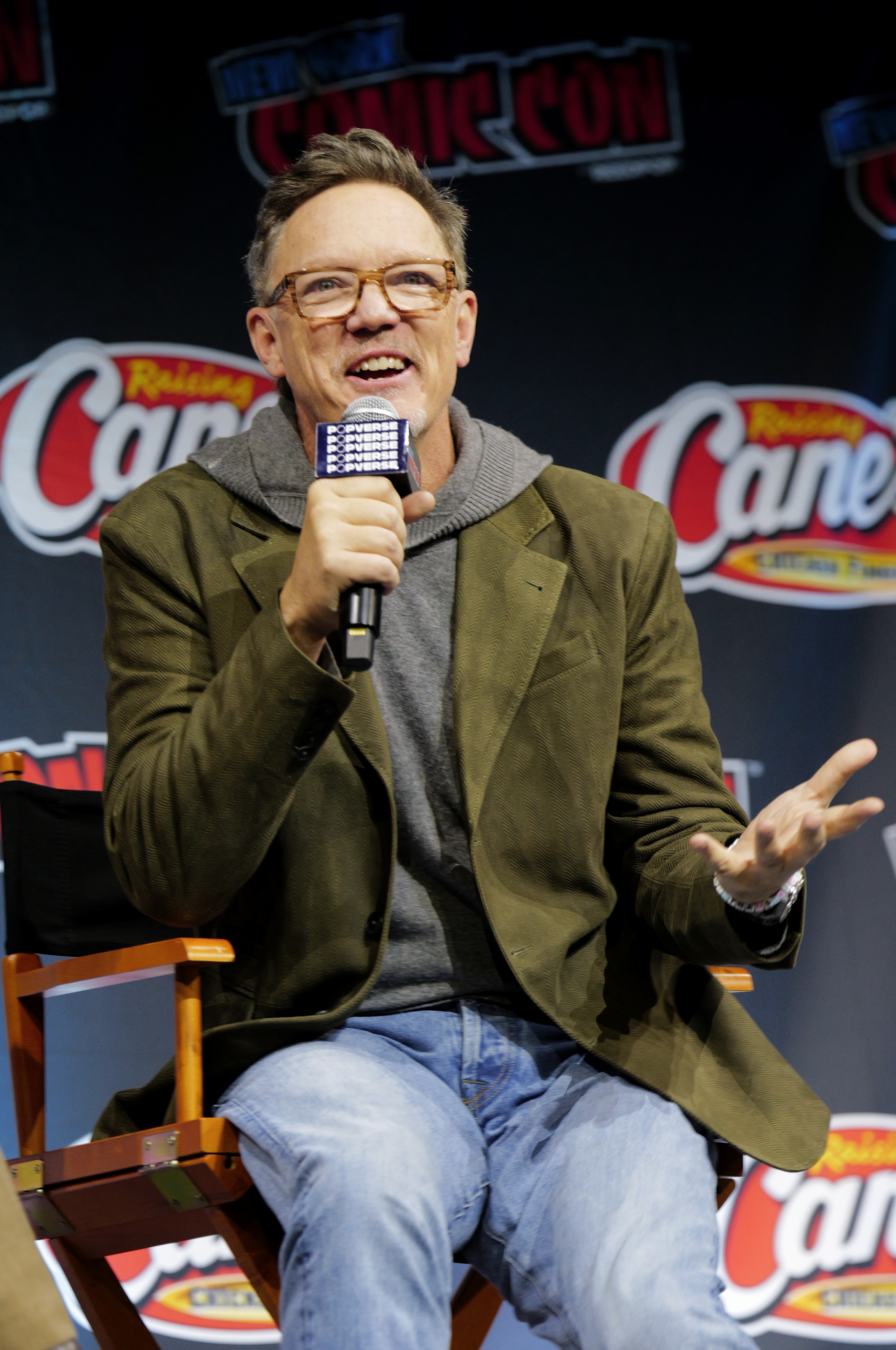 Matthew Lillard speaking on stage at a comic convention, wearing a blazer over a hoodie. They're seated on a director's chair, holding a microphone
