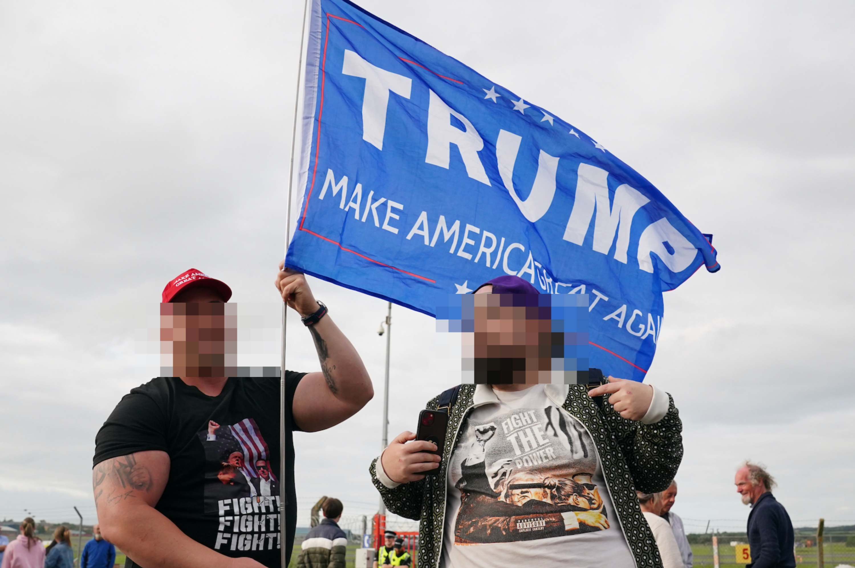 Two individuals hold a &quot;Make America Great Again&quot; flag at an outdoor event. One wears a cap and graphic T-shirt, and the other wears a T-shirt and patterned jacket