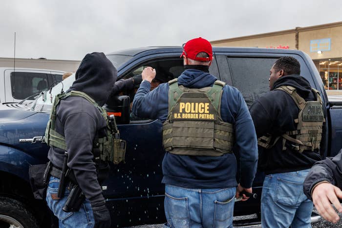 Three U.S. Border Patrol officers in tactical gear approach a dark truck, engaging with someone inside