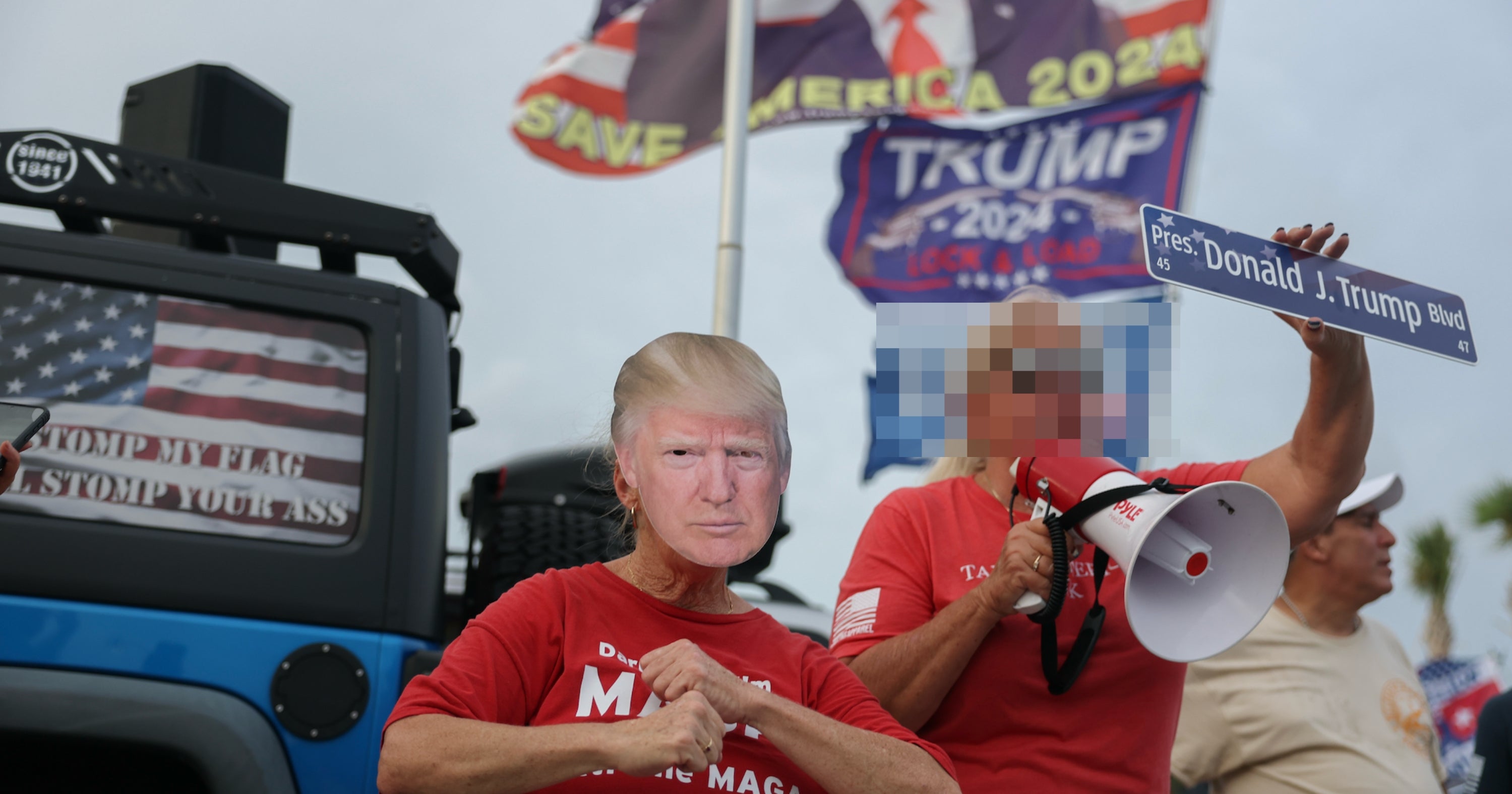 People at a rally: one in a Trump mask, another with a megaphone. Signs supporting Trump 2024 in the background