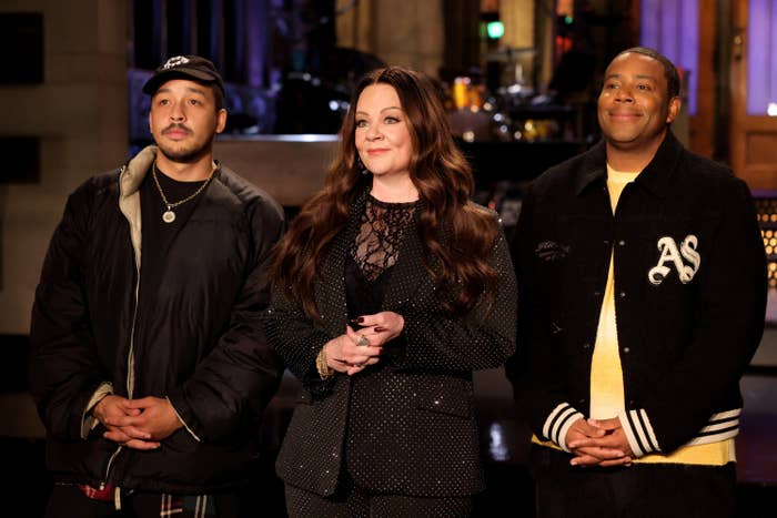 Three people on a stage, one in a lace outfit, stand side by side, smiling. Casual and patterned casualwear styles are worn by the two others