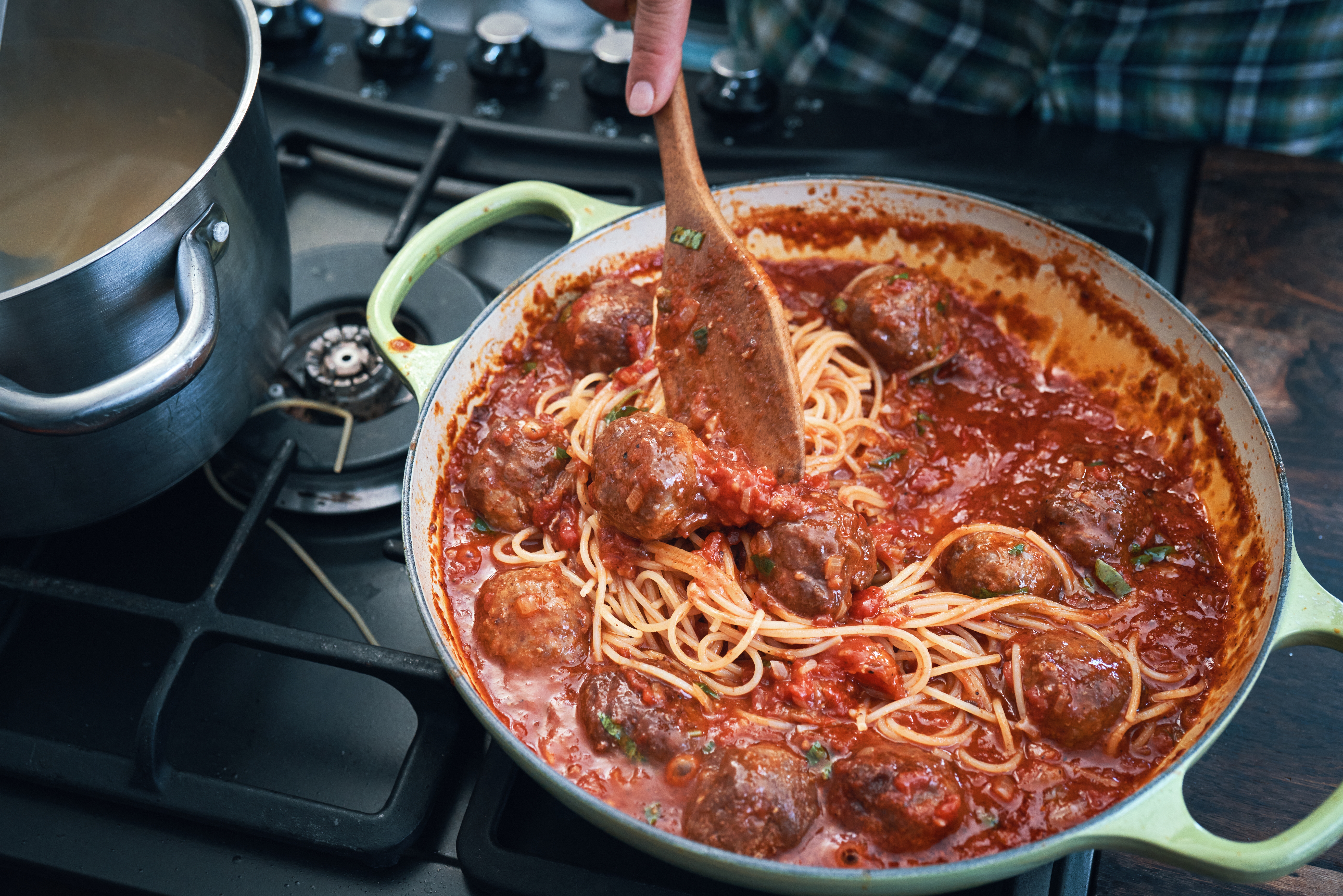 Person stirring spaghetti and meatballs in tomato sauce with a wooden spoon on a stove