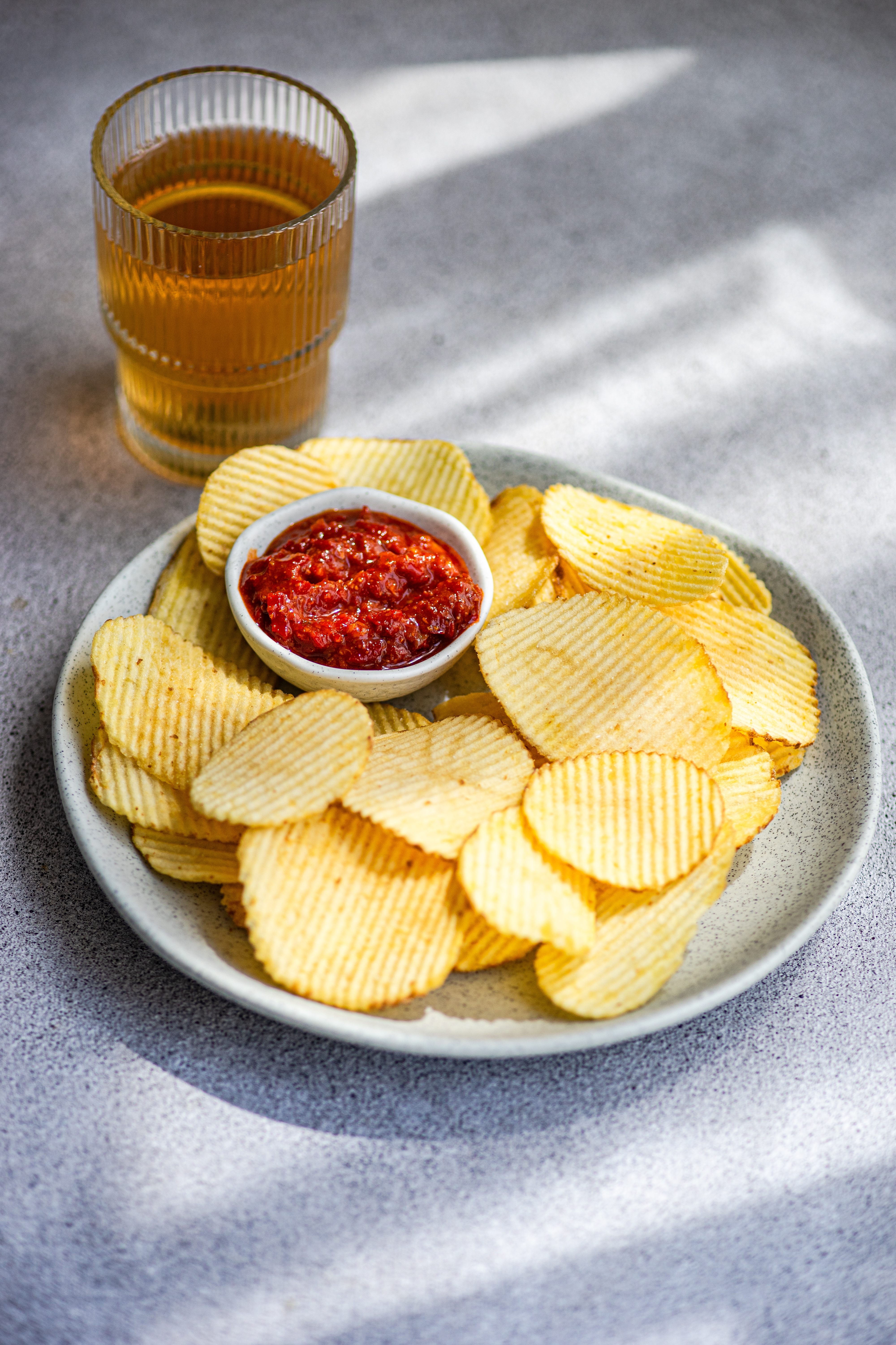 Plate of ridged potato chips with a small bowl of red salsa, alongside a glass of iced tea on a gray table