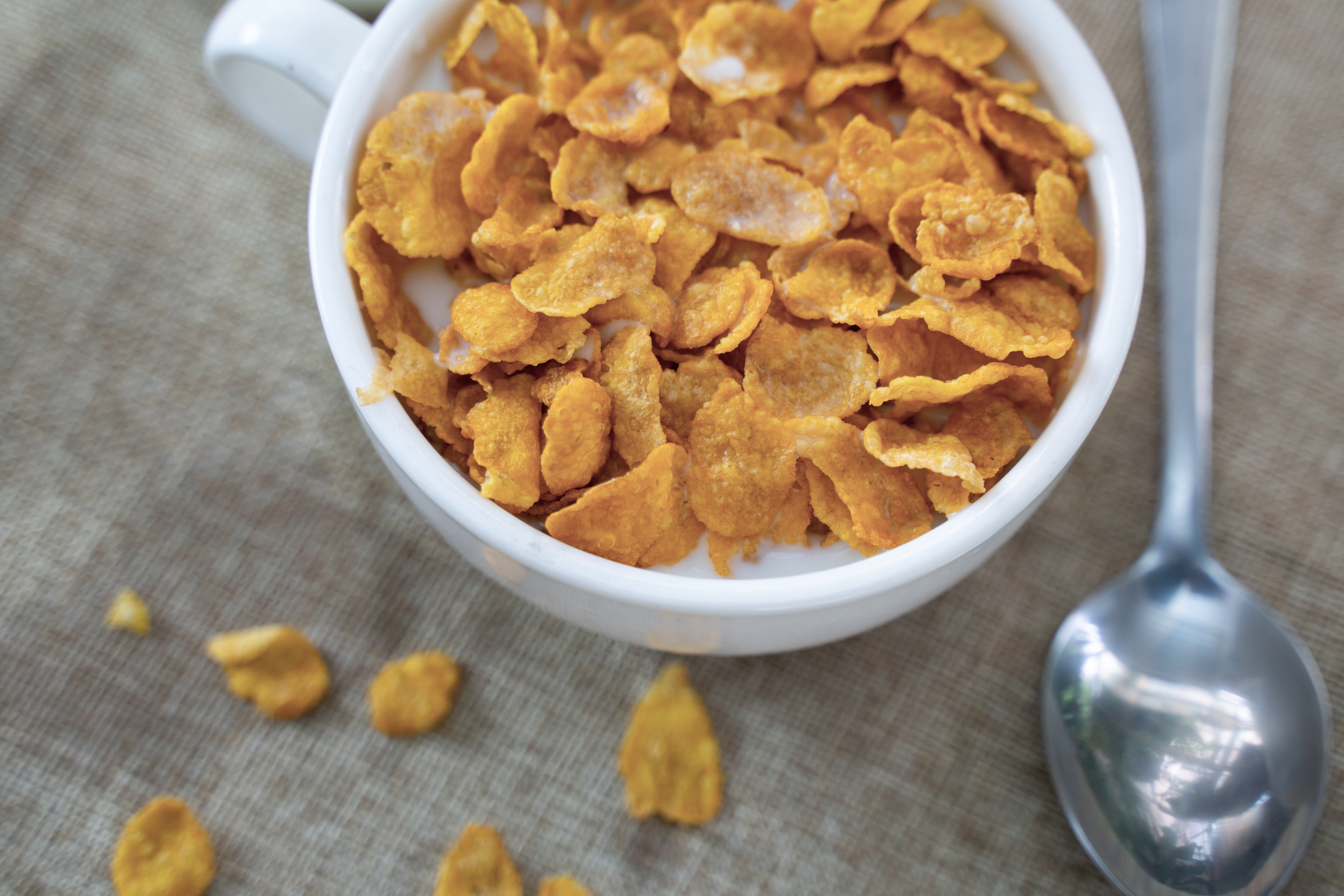 A bowl of cornflakes with milk, sitting on a burlap cloth, and a spoon beside it