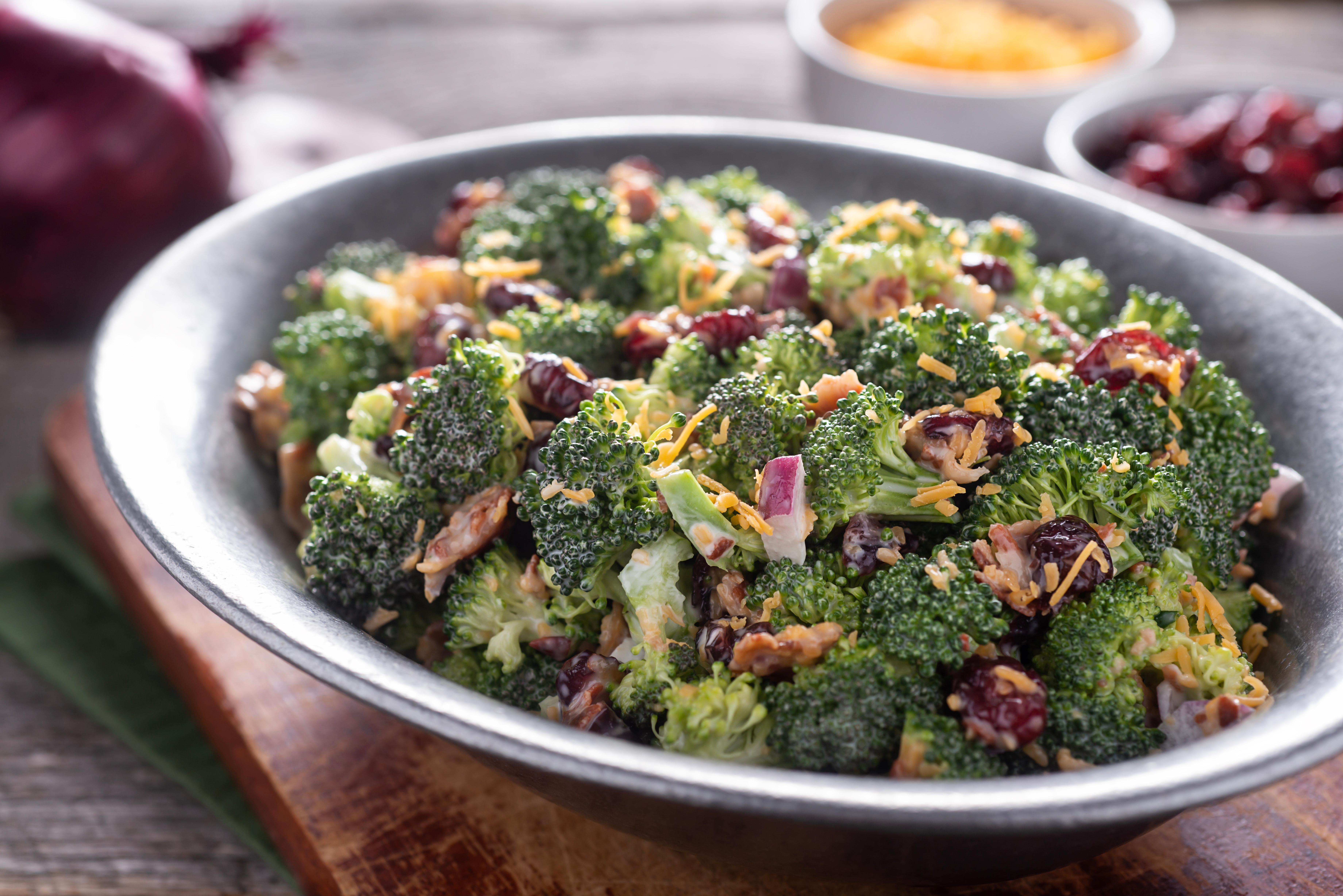 Broccoli salad with cranberries, walnuts, and a creamy dressing in a metal bowl on a wooden board, with toppings in small bowls nearby
