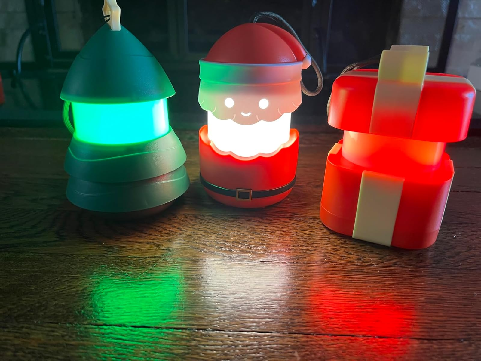 Three festive lanterns: a green Christmas tree, a Santa Claus, and a red gift box, illuminating a wooden surface