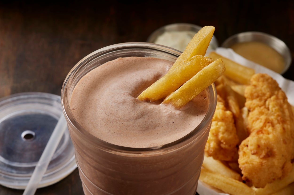 Chocolate milkshake with two fries inside, near fried chicken and more fries on a plate